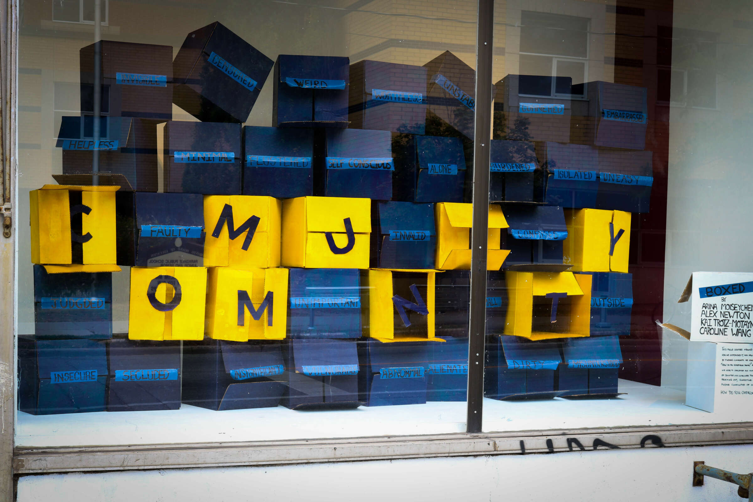 A window display features black and yellow boxes arranged in rows. The yellow boxes spell out "COMMUNITY" in a scattered manner, with a sign in the corner containing additional text that's not fully readable.