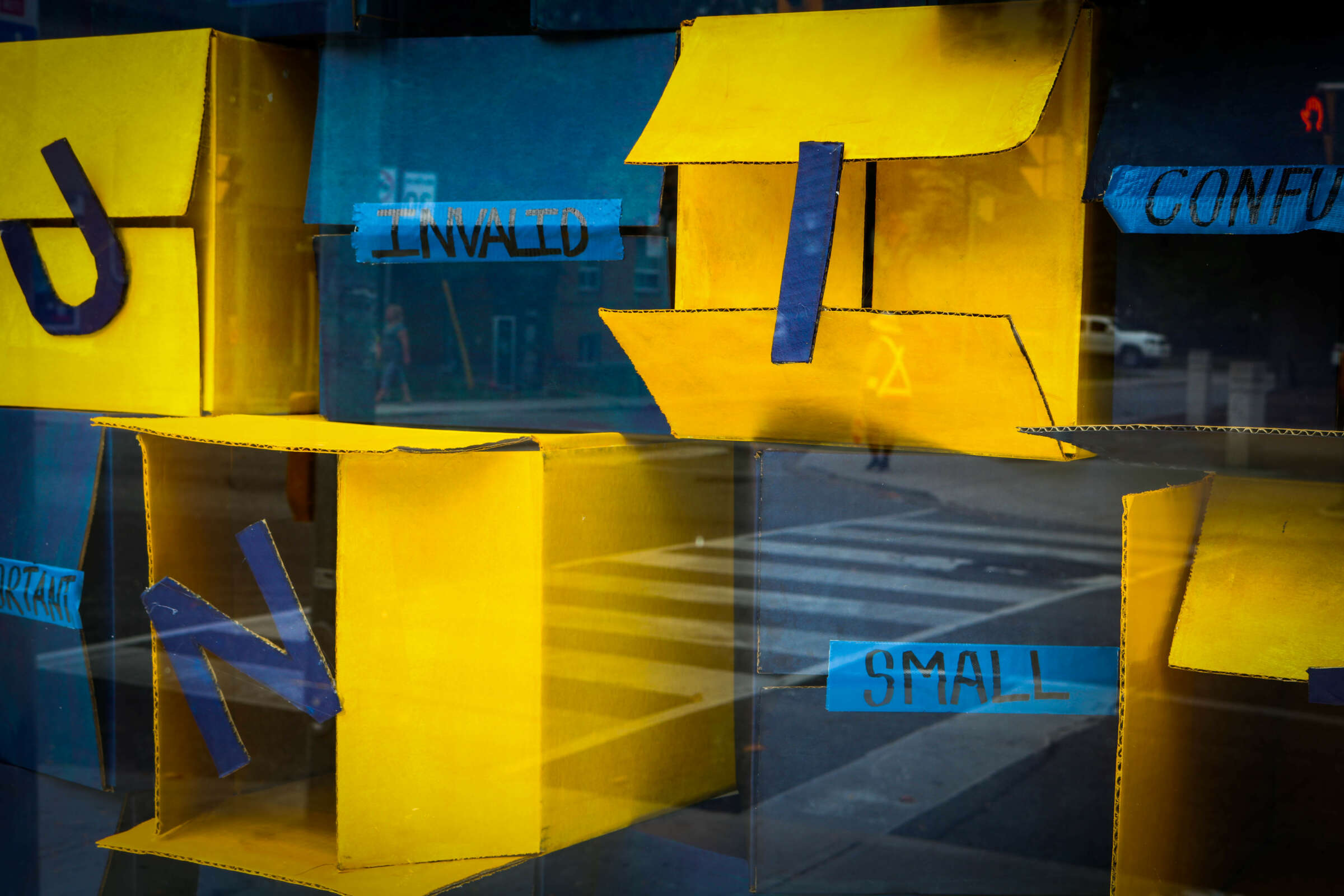 A window display featuring yellow, open cardboard boxes with blue tape forming letters "N," "U," and "I" on them. Blue paper labels with words like "INVALID," "CONFUSED," and "SMALL" are attached to the boxes. A street and crosswalk are visible outside.