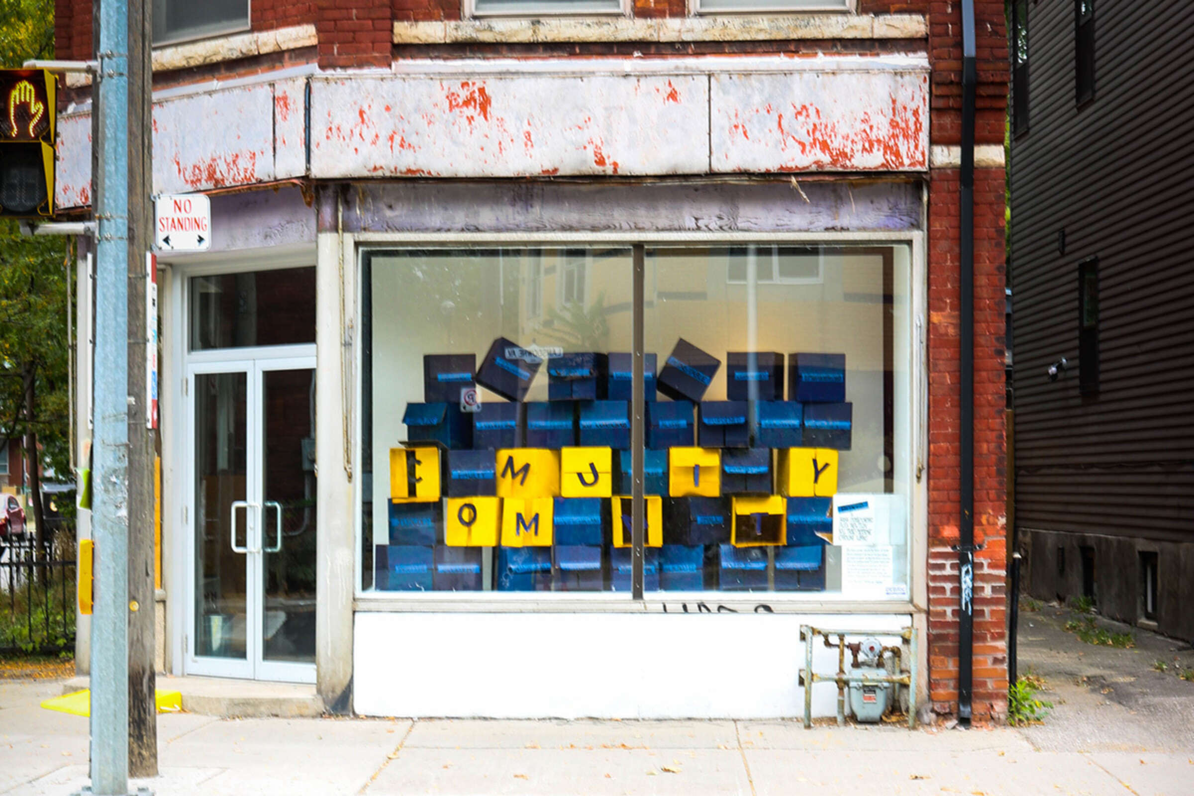 A storefront with large windows displays abstract blue paintings. Yellow letters near the bottom of the windows appear to spell out "FAMILY" among the art. The surrounding red brick building shows signs of weathering, and a "No Standing" sign is visible on the sidewalk.