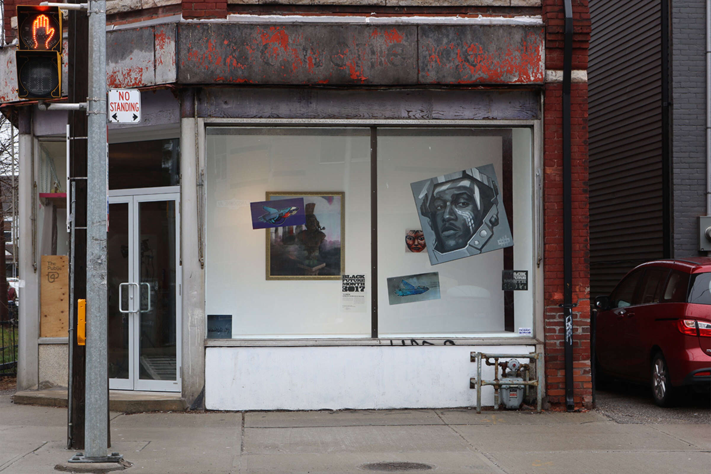 A street view of an art gallery window in a weathered brick building. The display features various paintings, including a large grayscale portrait and colorful abstract works. There's a red minivan parked nearby and a "No Standing" traffic sign visible.