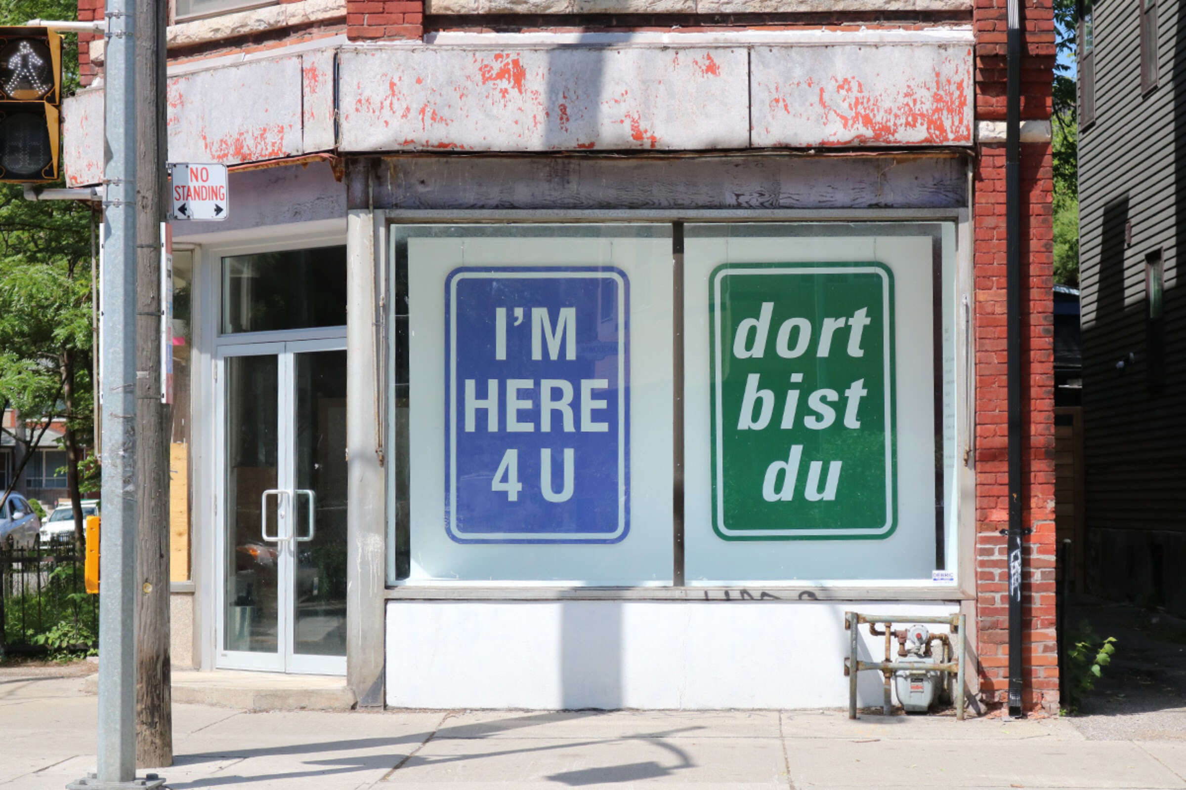 A street view of a building corner with windows displaying two large signs. The left sign is blue with white text saying "I'M HERE 4 U," and the right sign is green with white text saying "dort bist du." The building's exterior appears slightly worn.