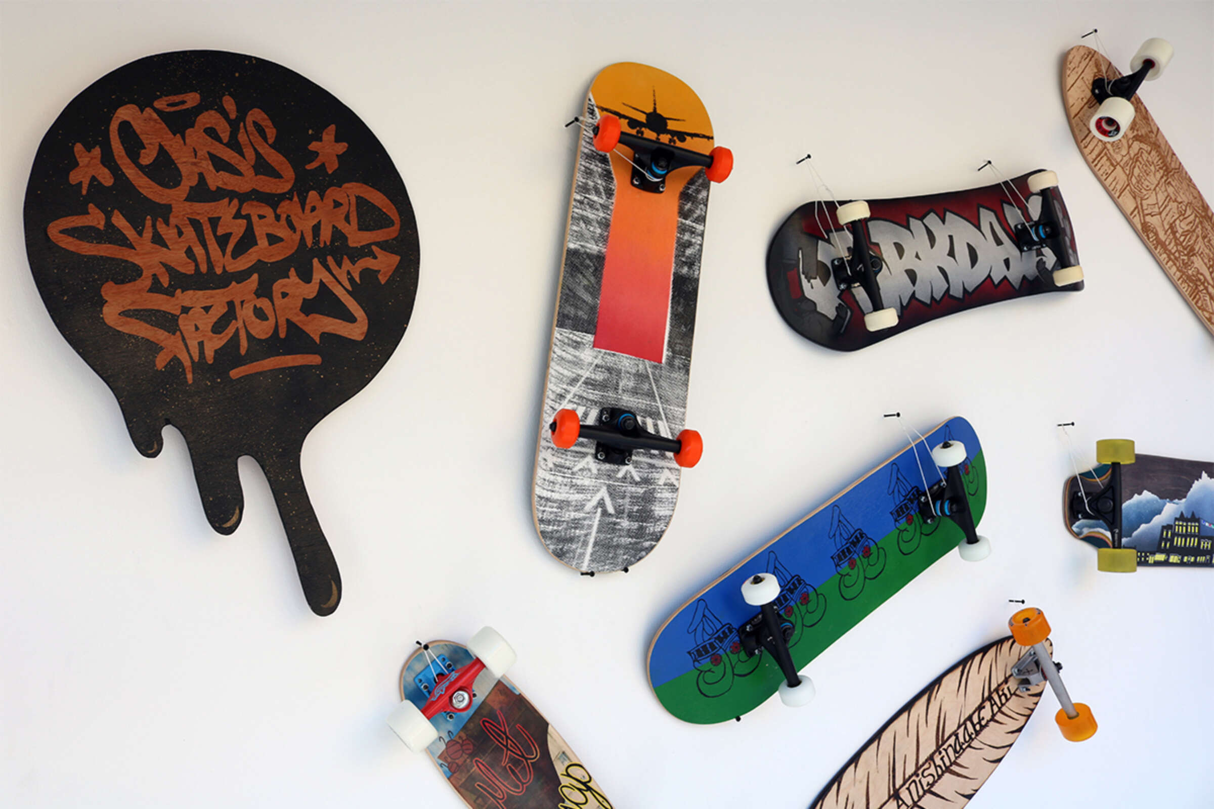 A white wall with various skateboards mounted on it. The skateboards have unique designs, including graffiti, landscapes, and abstract artwork. A sign shaped like a melting splash reads "Classic Skateboard Factory" in graffiti-style text.