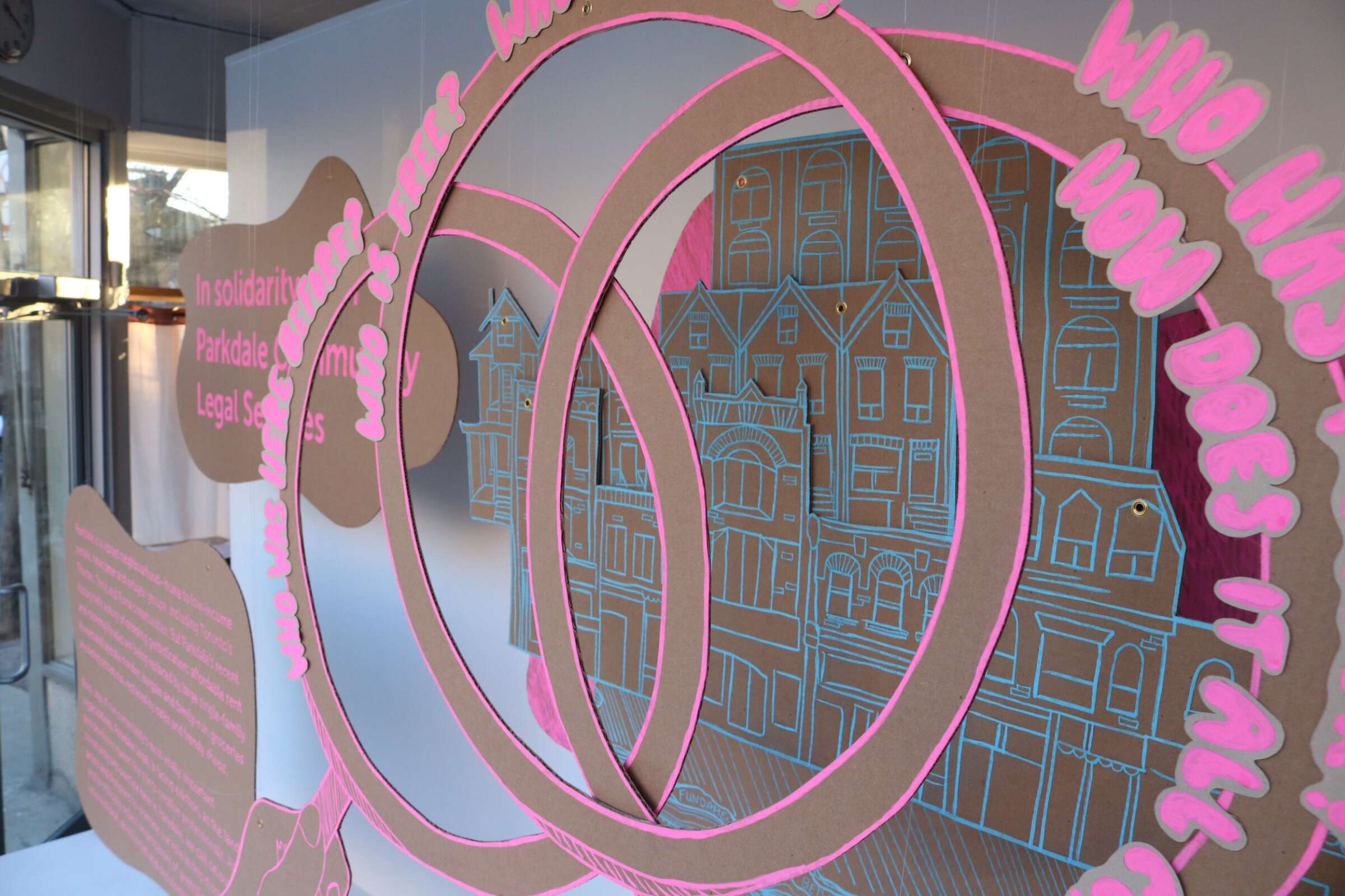 An intricate artwork features three large concentric rings with illustrations of buildings and text. Bright pink outlines highlight the cardboard structures. The phrases "In solidarity with Parkdale's Legal Services" and "Who has the right? Who does it harm?" are visible.