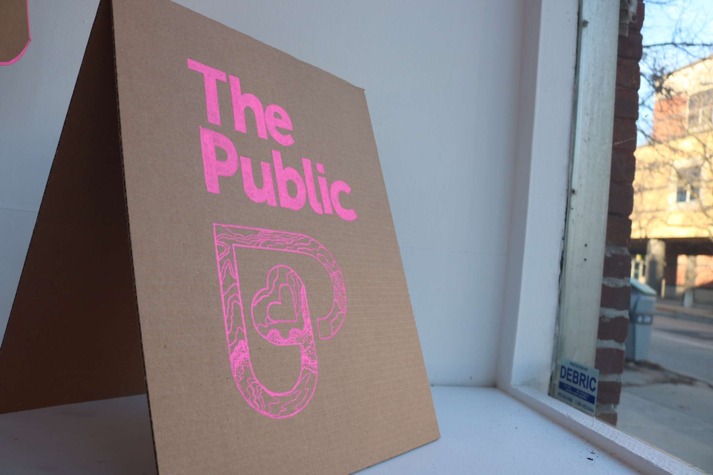 A cardboard sign with the text "The Public" and a graphic of a stylized heart in pink, displayed indoors near a window. Outside, a street scene with brick buildings and a tree is visible in the background.