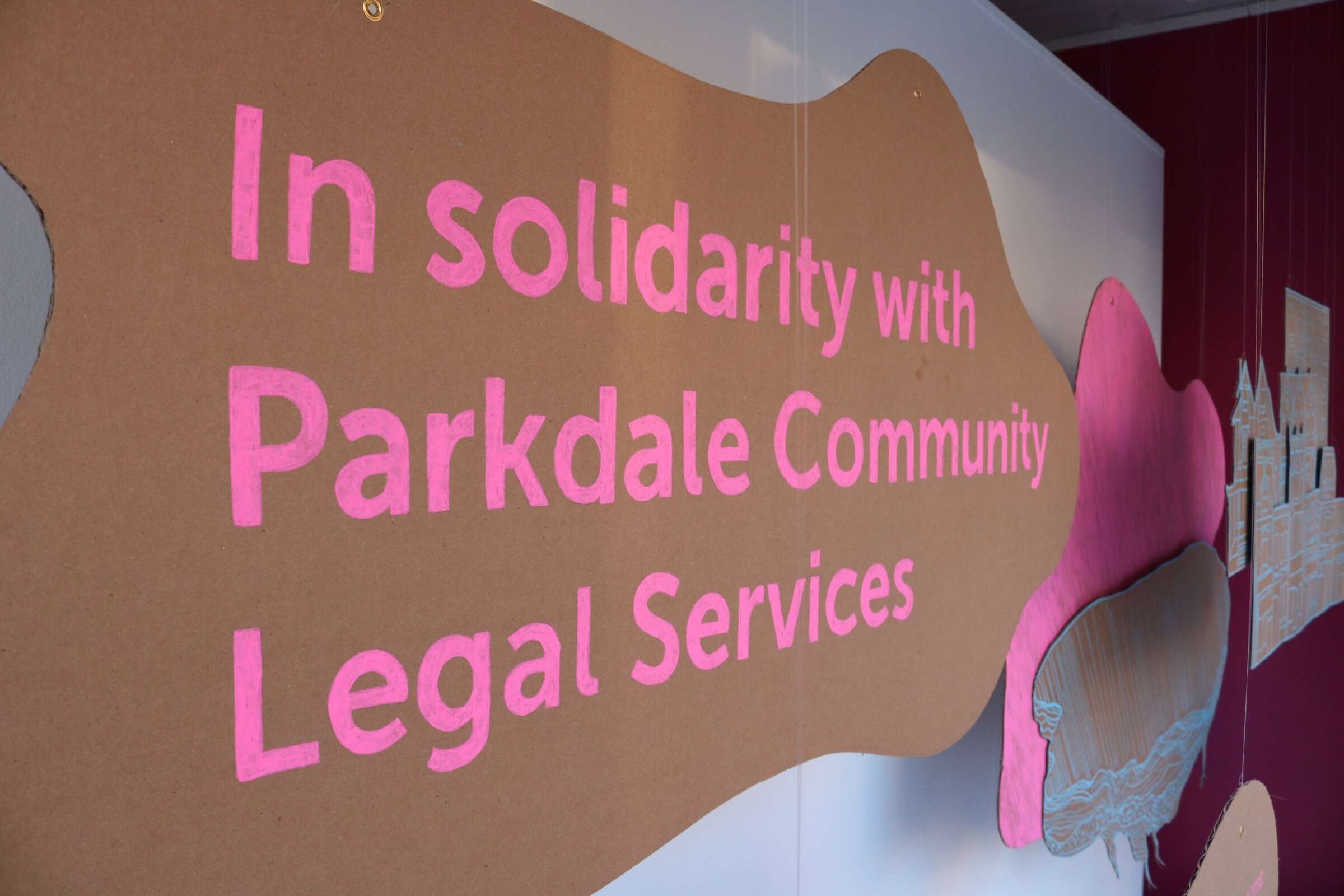 A cardboard sign with pink lettering reads "In solidarity with Parkdale Community Legal Services." The sign is displayed against a white wall with a pink abstract shape in the background.