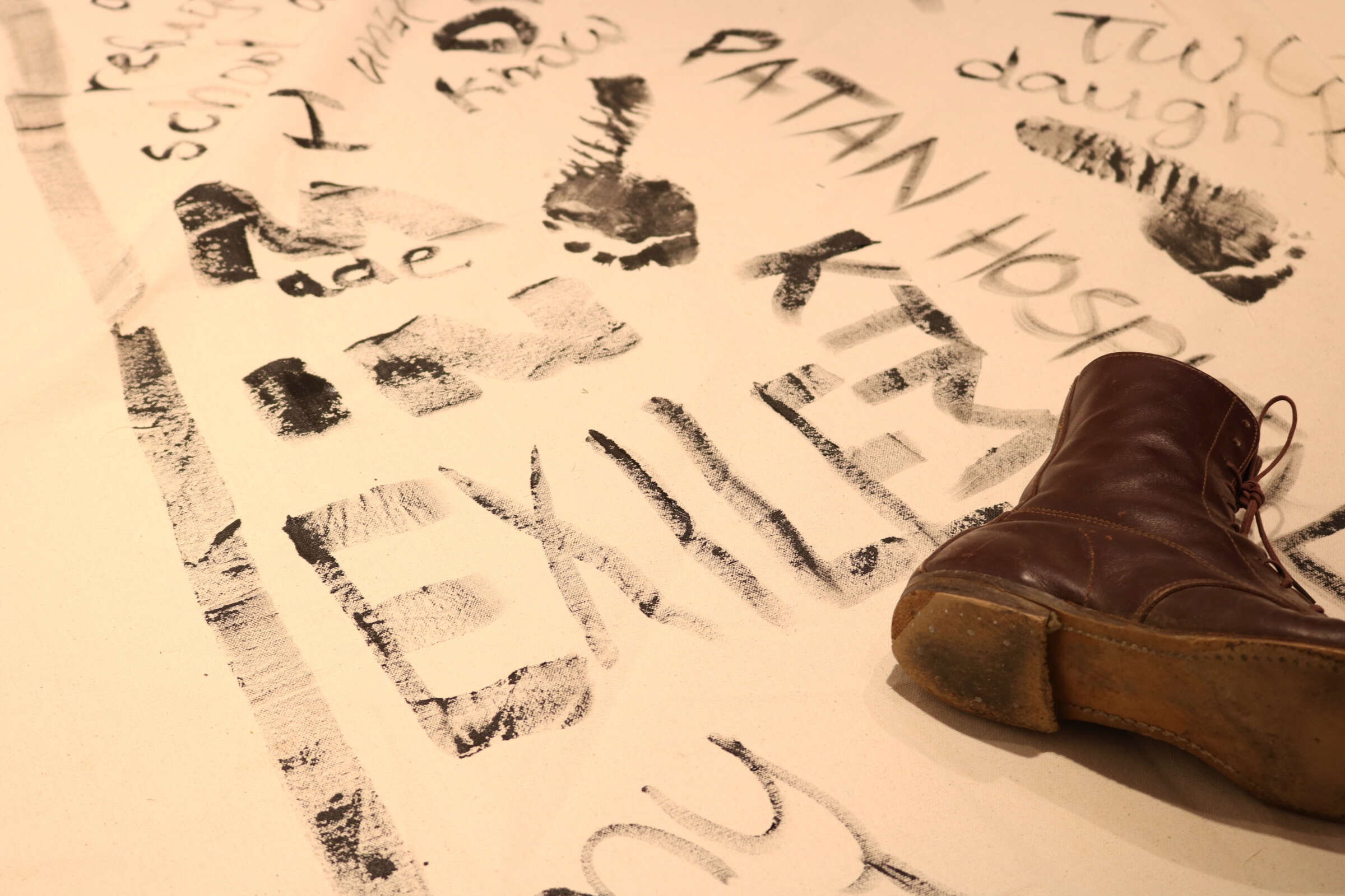 An art piece featuring a brown boot and various smeared black ink writings and imprints on a beige surface. The word "EXILES" is prominently visible. Other partial words and phrases, footprints, and smudges create a chaotic backdrop.