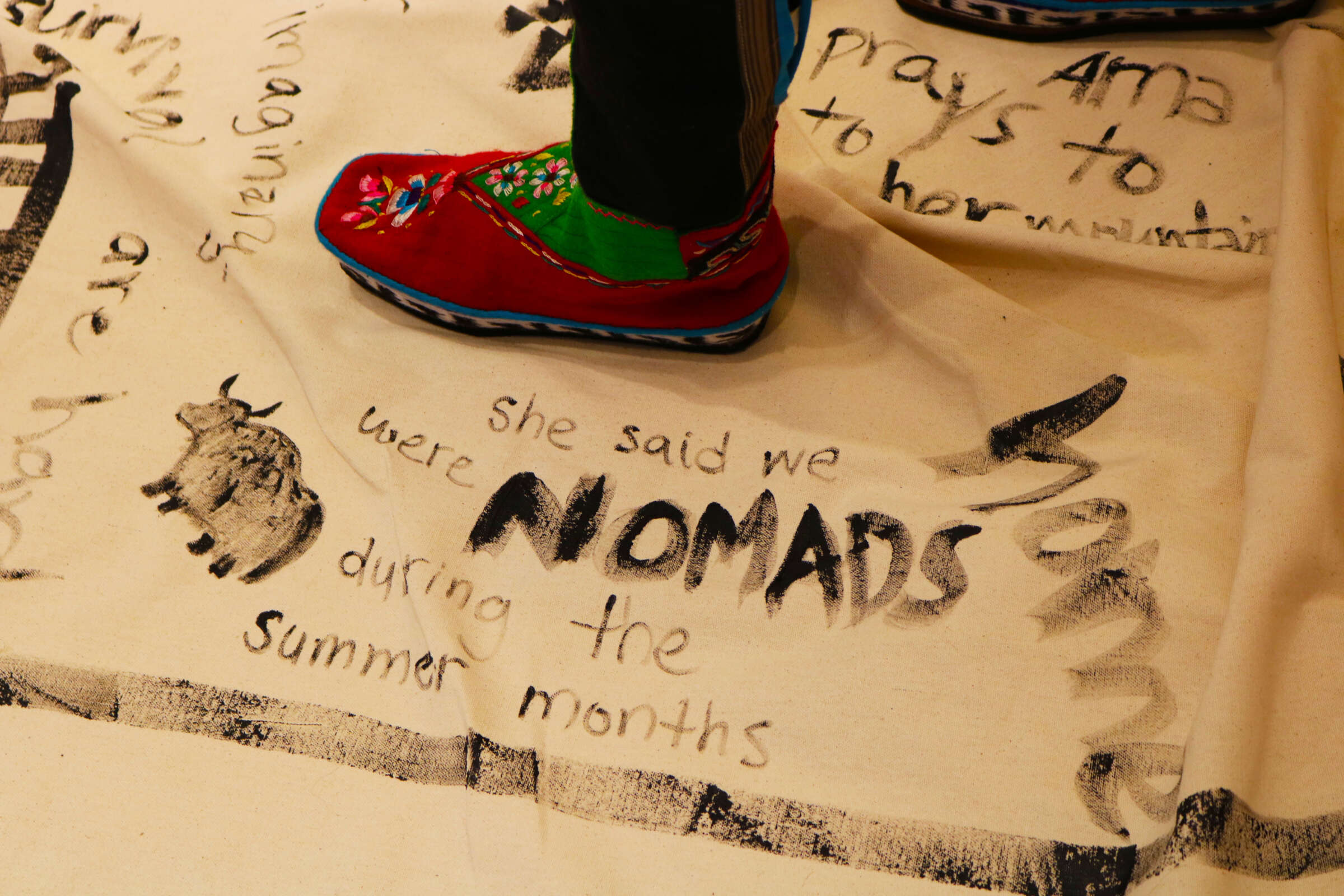 A close-up of colorful moccasin-clad feet standing on a cream-colored cloth. The cloth features handwritten text and drawings in black ink, including the words "she said we were NOMADS during the summer months" and an illustration of an animal.