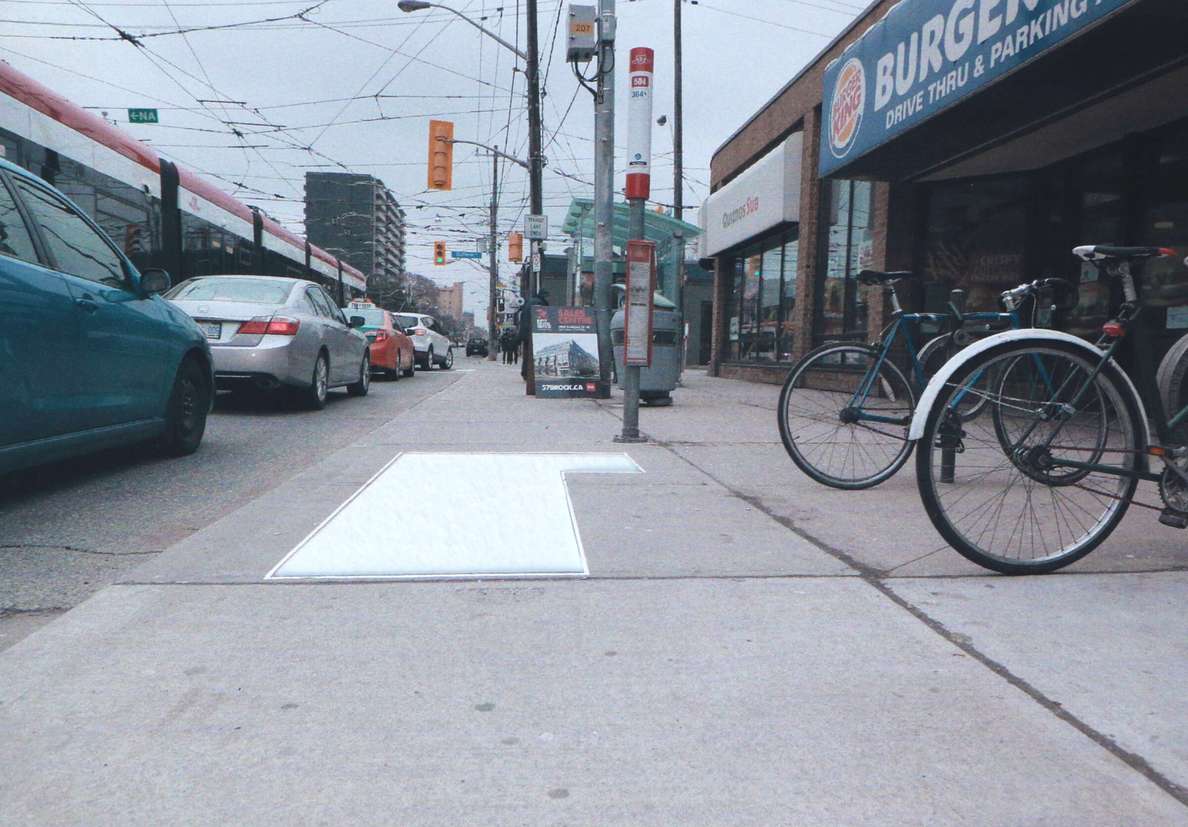 A city street scene with several parked cars and a light rail train in the background. The sidewalk has a white painted arrow, a parked bicycle, and nearby is a Burger Joint with a "Drive Thru & Parking" sign. Overhead, there are numerous power lines.