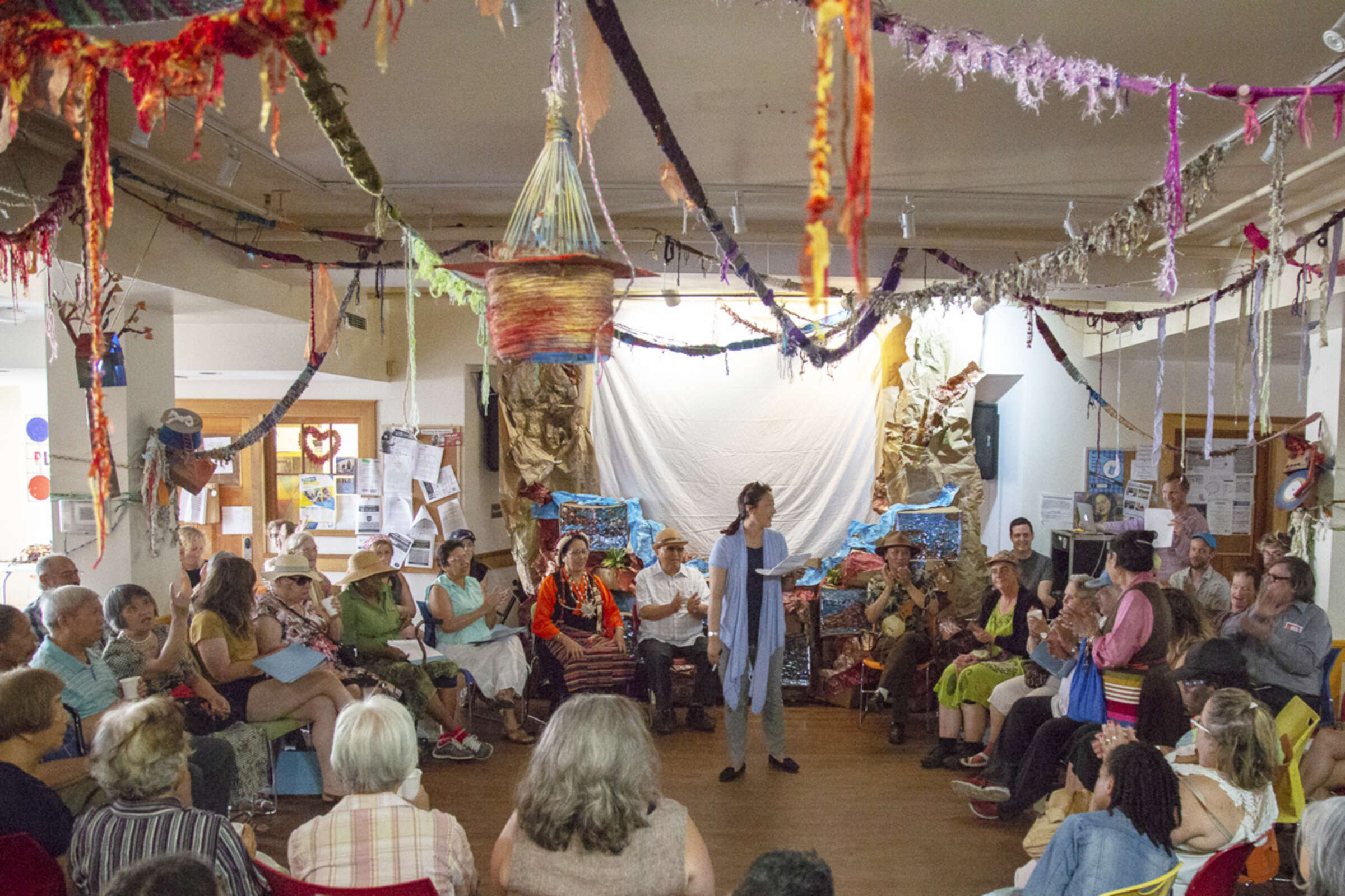 A large group of people sit in a circle in an eclectic room decorated with colorful yarn art hanging from the ceiling. A person stands in the center, speaking, while others listen attentively. The setting appears casual and artistic, with various decorations adorning the space.