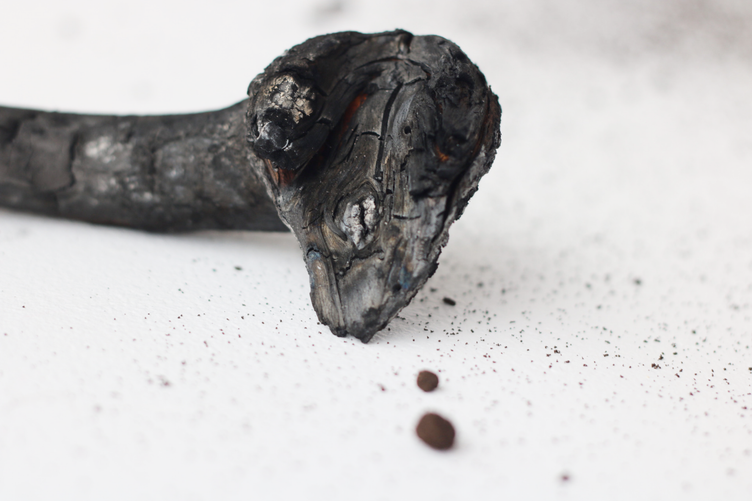 Close-up of a blackened, charred piece of wood shaped like a bent rod resting on a white surface with scattered ash and soot particles around it. The focus is on the textured, burned end of the wood, highlighting the intricate details of the charred surface.