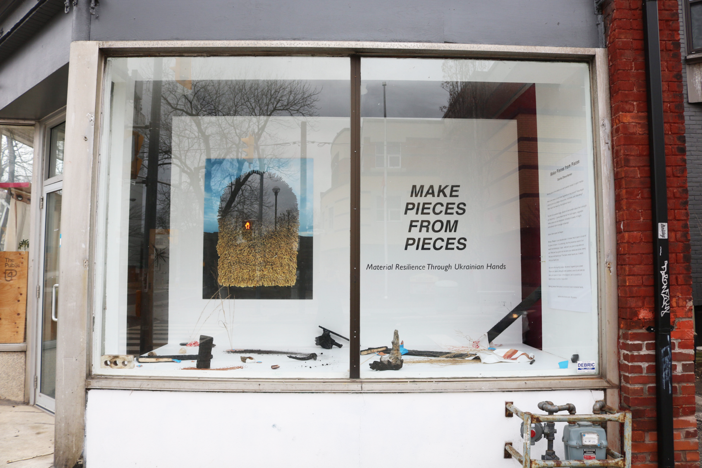 A storefront display features an art exhibition titled "MAKE PIECES FROM PIECES: Material Resilience Through Ukrainian Hands." The window showcases various art pieces, including mixed media and sculptures, set against a white backdrop with large text.