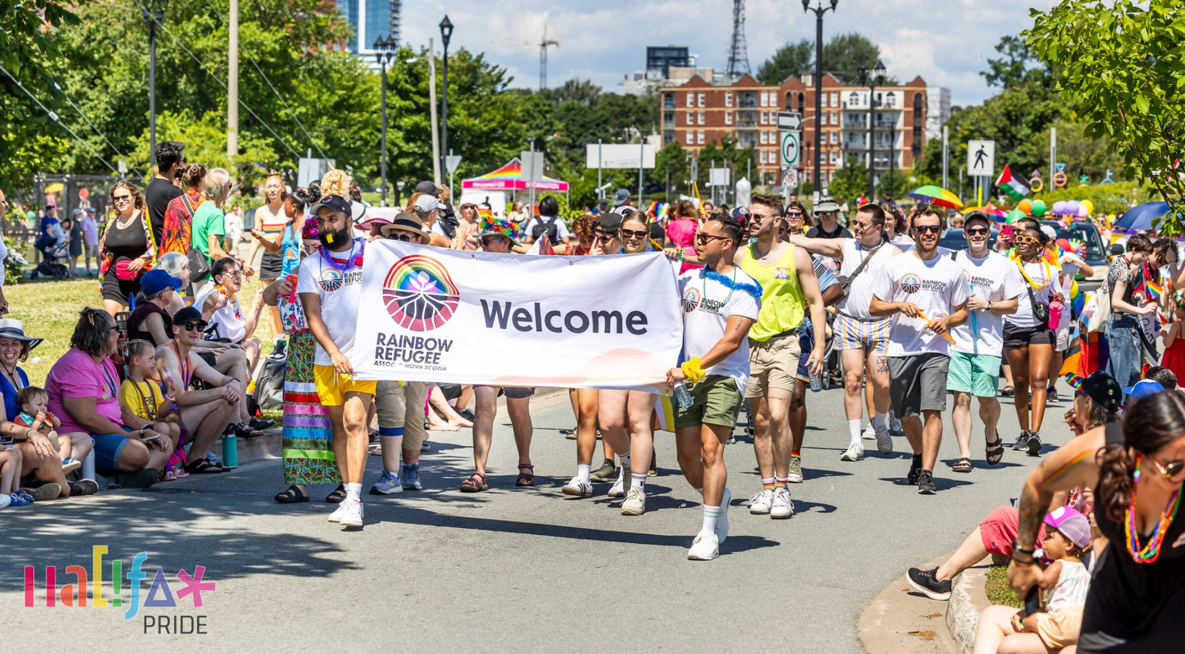 A group of people proudly march down a street holding a large sign that reads "Welcome Rainbow Refugee." The event is festive, with colorful outfits and flags. Spectators line the street, some seated and others standing, enjoying the parade. The "Inla Pride" logo is visible in the bottom left corner.