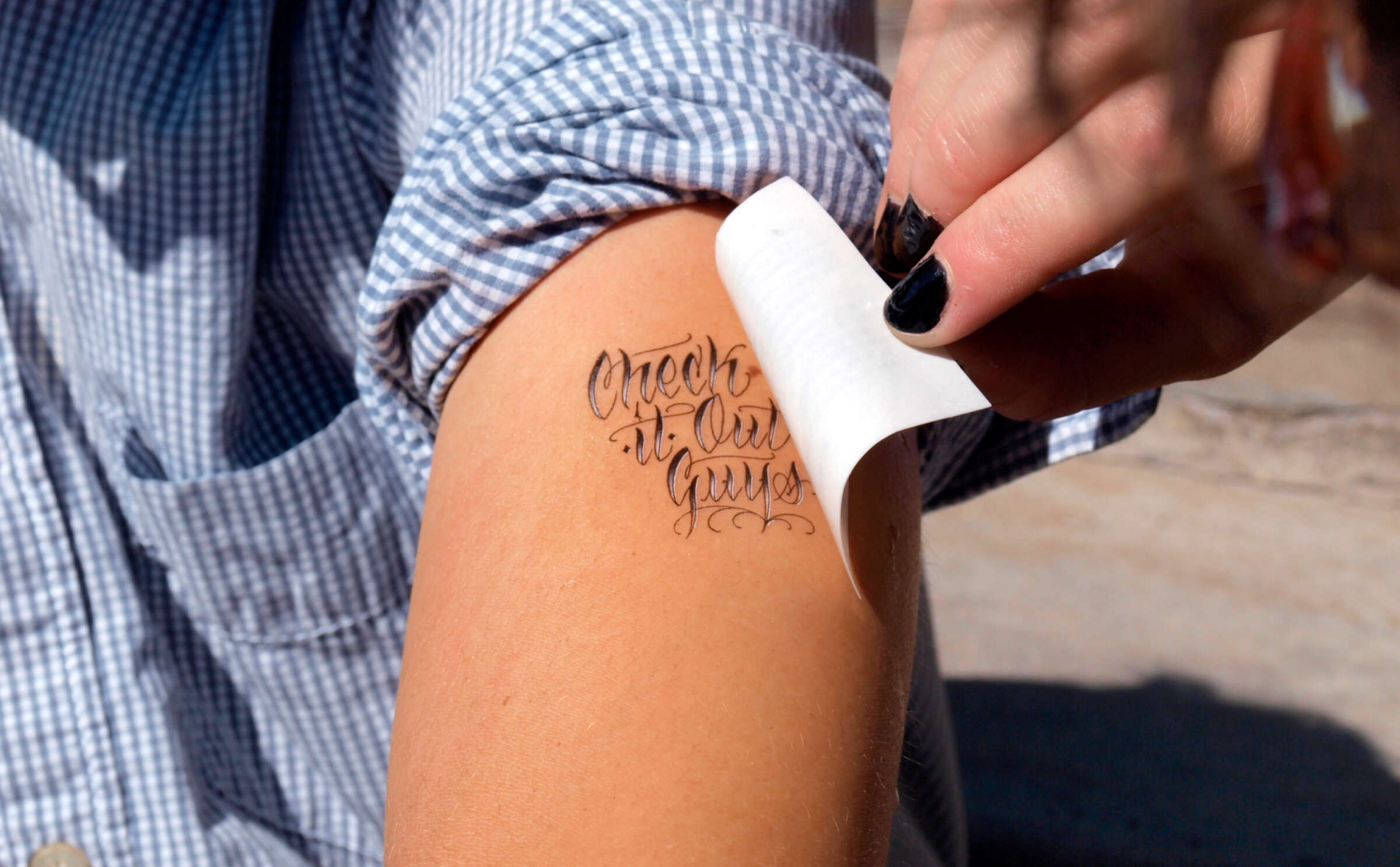 A person in a checkered shirt applies a temporary tattoo on their arm that reads "Check It Out Guys" in cursive script. Their nails are painted black.