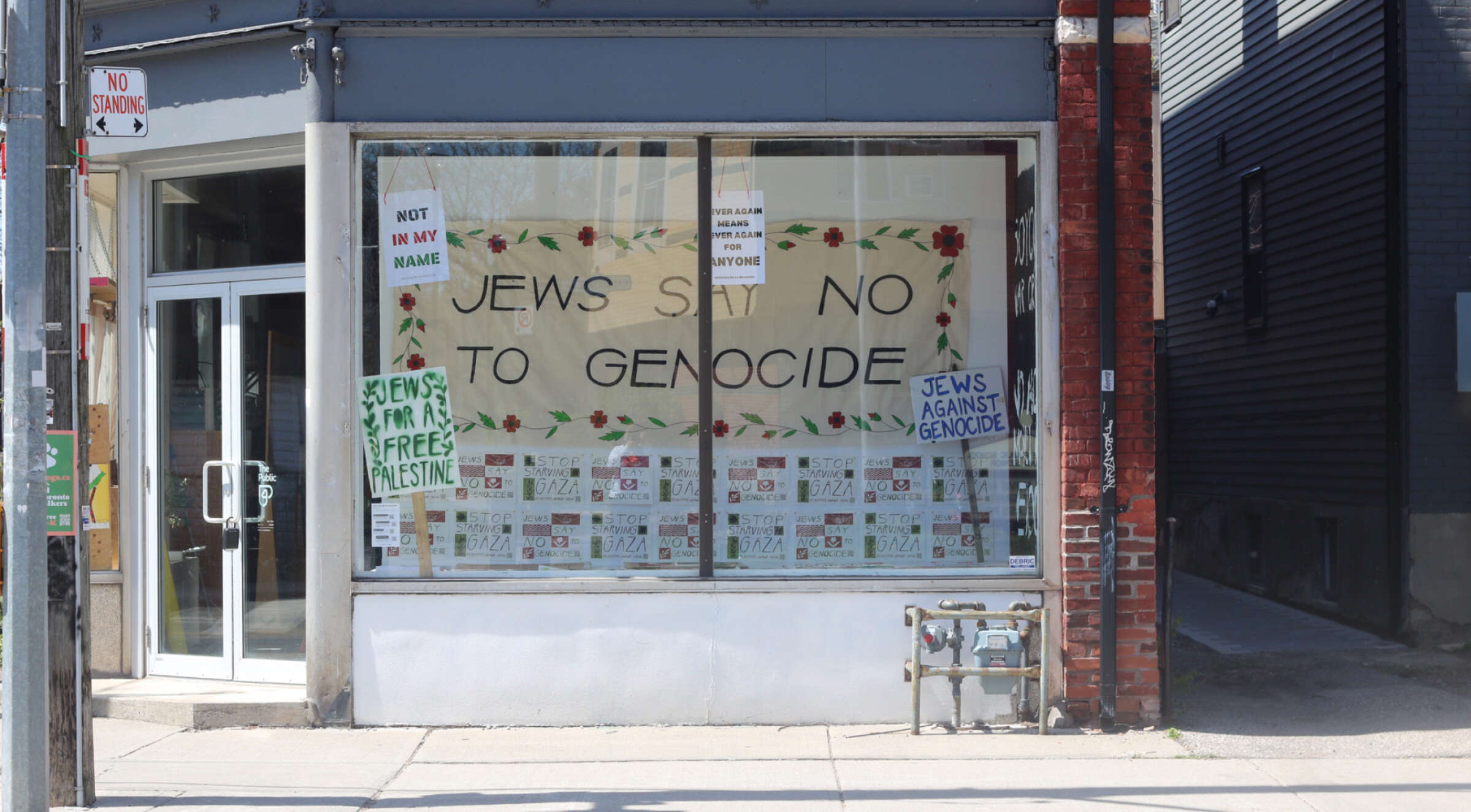 A storefront window displays a large sign that reads "Jews Say No to Genocide" with smaller posters that say "Not in my name" and other similar messages on both the window and the door, framed by a brick and metal facade. The posters support Palestinian freedom and oppose genocide.