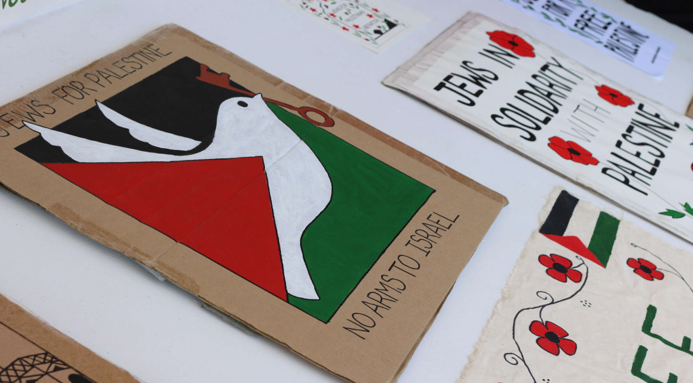 Various protest signs supporting Palestine lay on a flat surface. One prominent sign features a white dove over a stylized red, green, and black background and reads, "No Arms to Israel." Another nearby sign states, "Jews in Solidarity with Palestine.