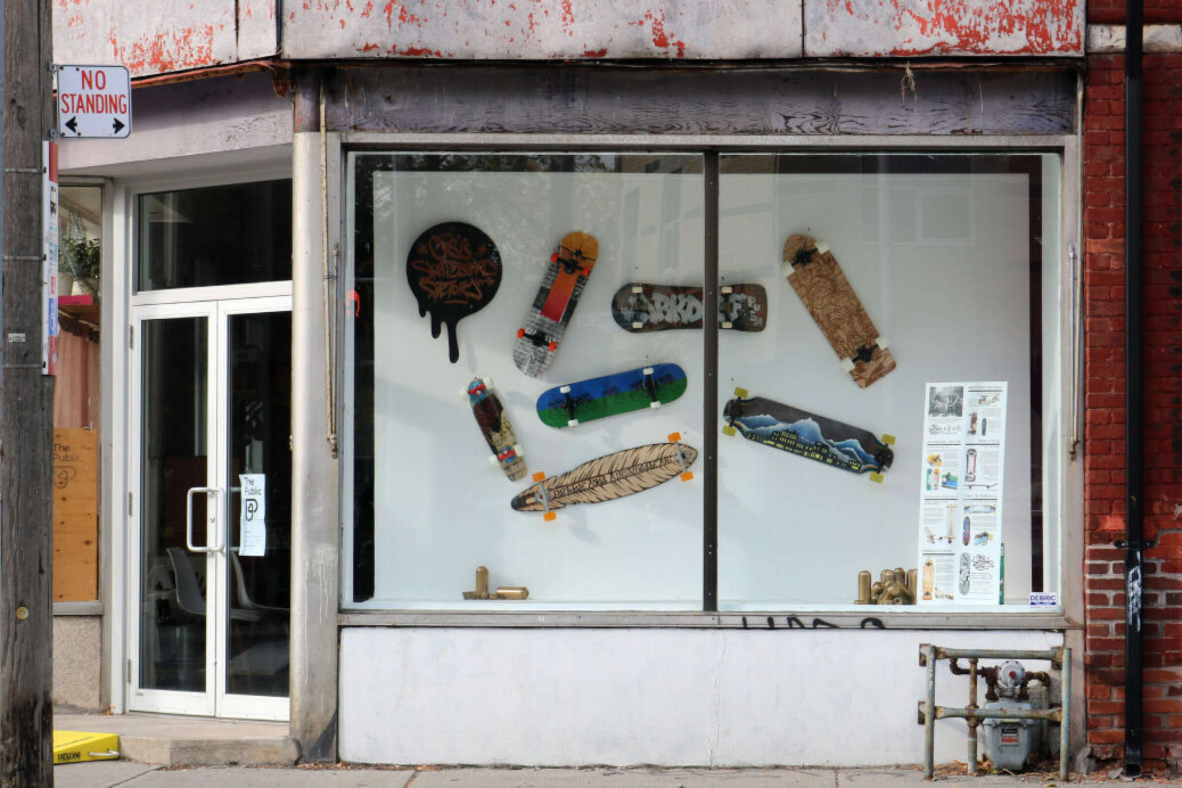 A storefront window display showcases several creatively designed skateboards against a plain white background. The skateboards exhibit various artistic designs, including painted graphics, a feather, and fur. A "No Standing" sign is visible on the left side of the storefront.