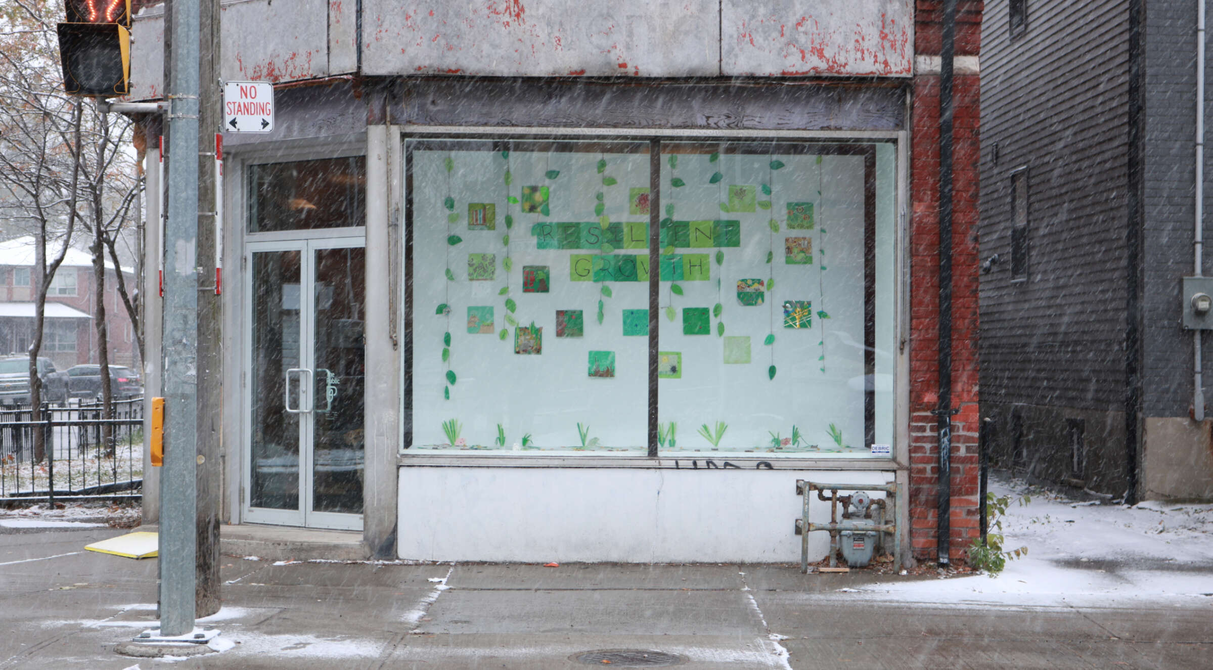 A street view of a corner building during snowfall. The building has a large window display featuring various green and yellow paper cutouts arranged on the glass. A "No Parking" sign is visible on a pole, and the ground is lightly dusted with snow.