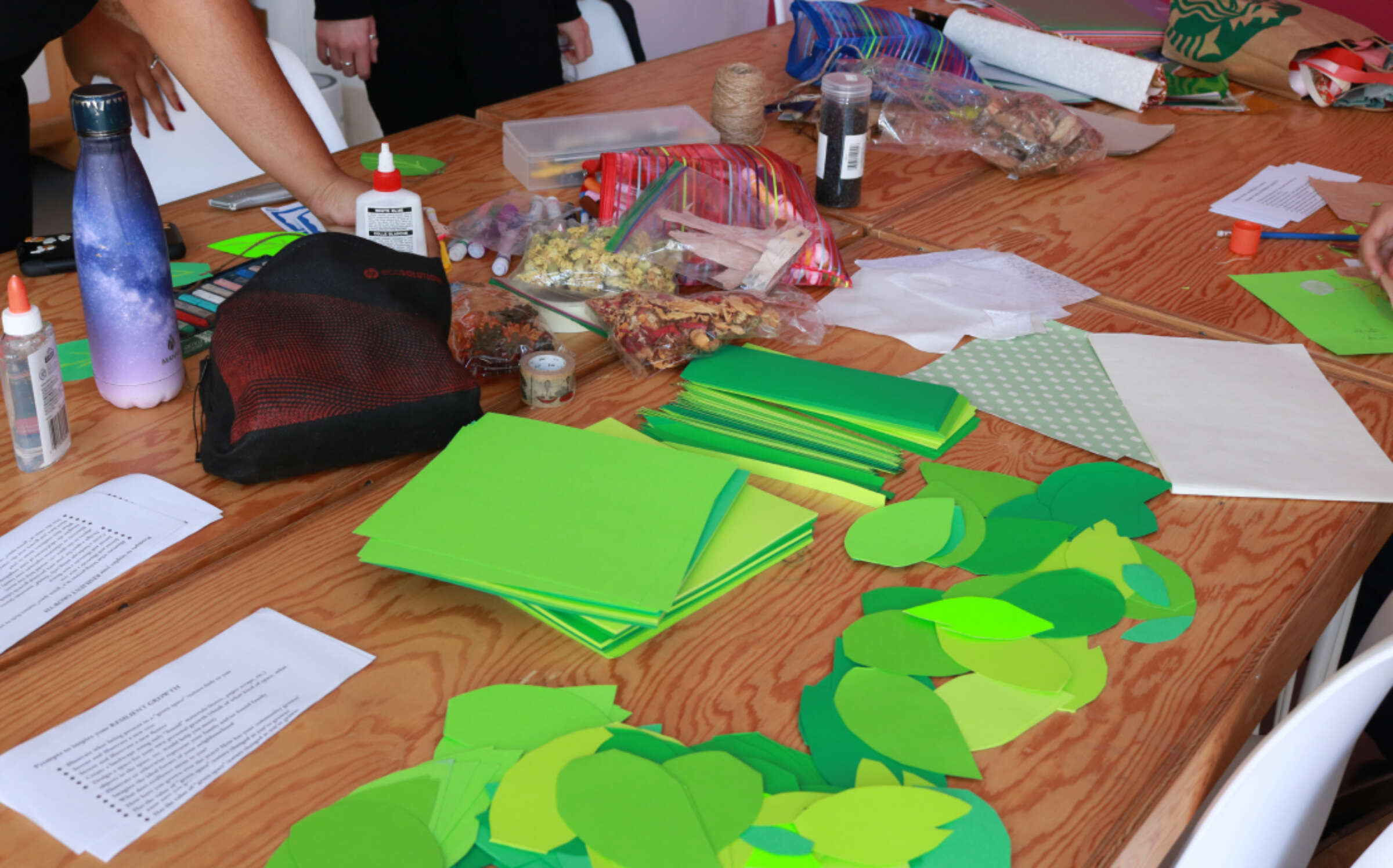 A wooden table covered with various crafting supplies, including green paper leaves, colored paper, glue bottles, scissors, markers, and a water bottle. There are two people partially visible, working on their projects.