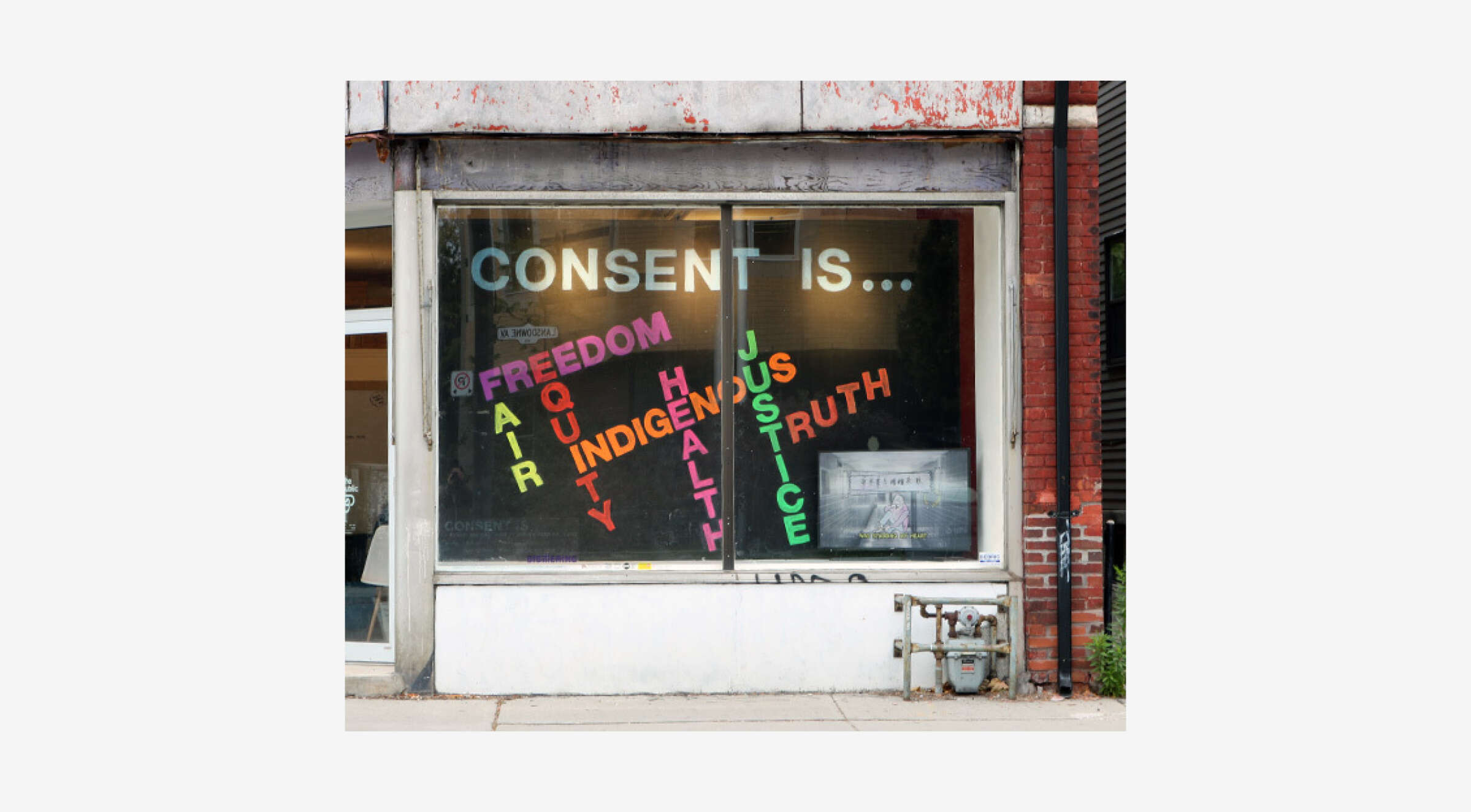 Storefront window with a large sign reading "CONSENT IS..." followed by colorful, vertically oriented words "FREEDOM, EQUITY, INDIGENOUS, HEALTH, JUSTICE, TRUTH." The backdrop shows a weathered building exterior with a smaller sign inside the window.