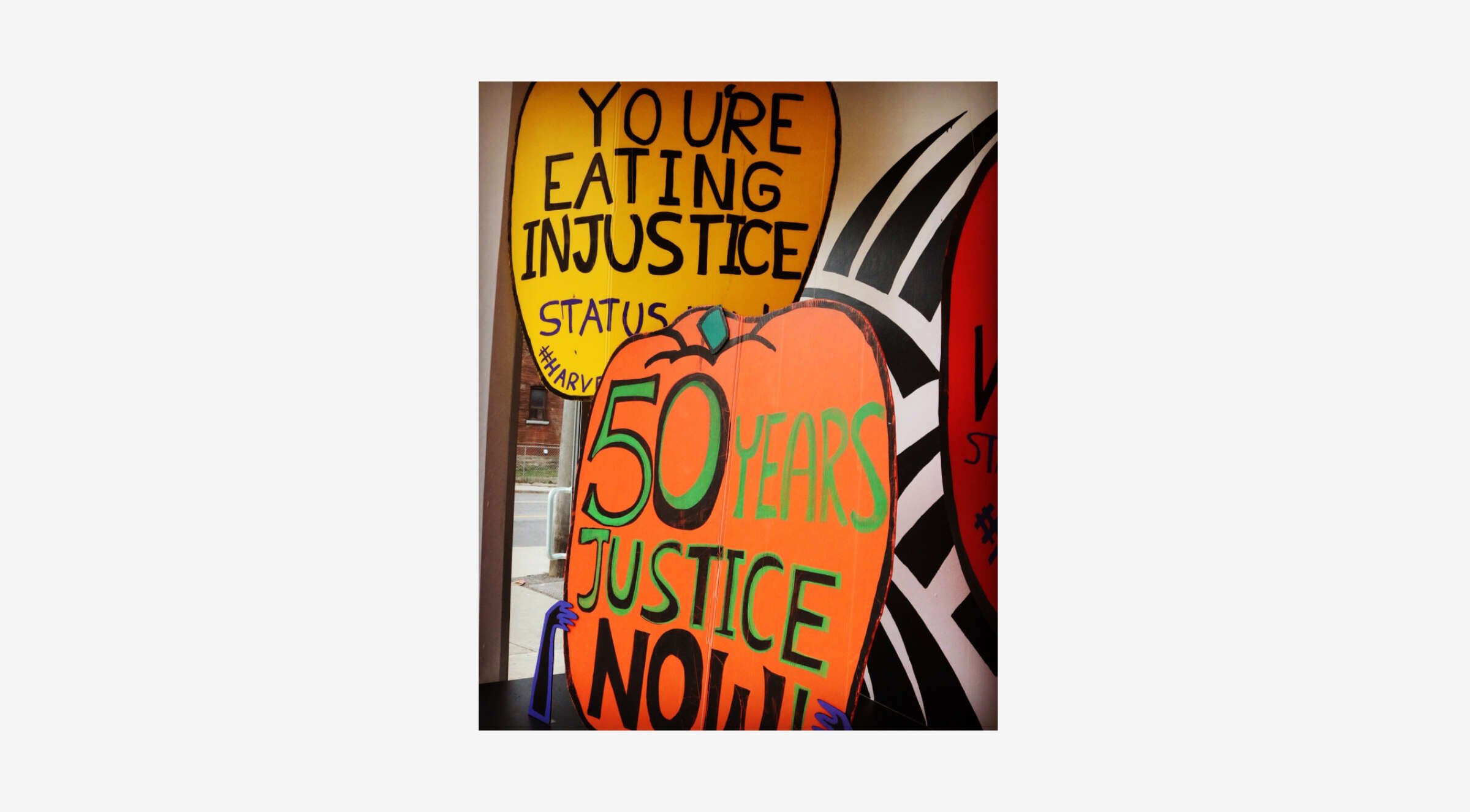 Brightly colored protest signs with messages like "YOU'RE EATING INJUSTICE" on a yellow balloon shape and "50 YEARS JUSTICE NOW!" on an orange heart shape are displayed against a street scene backdrop.