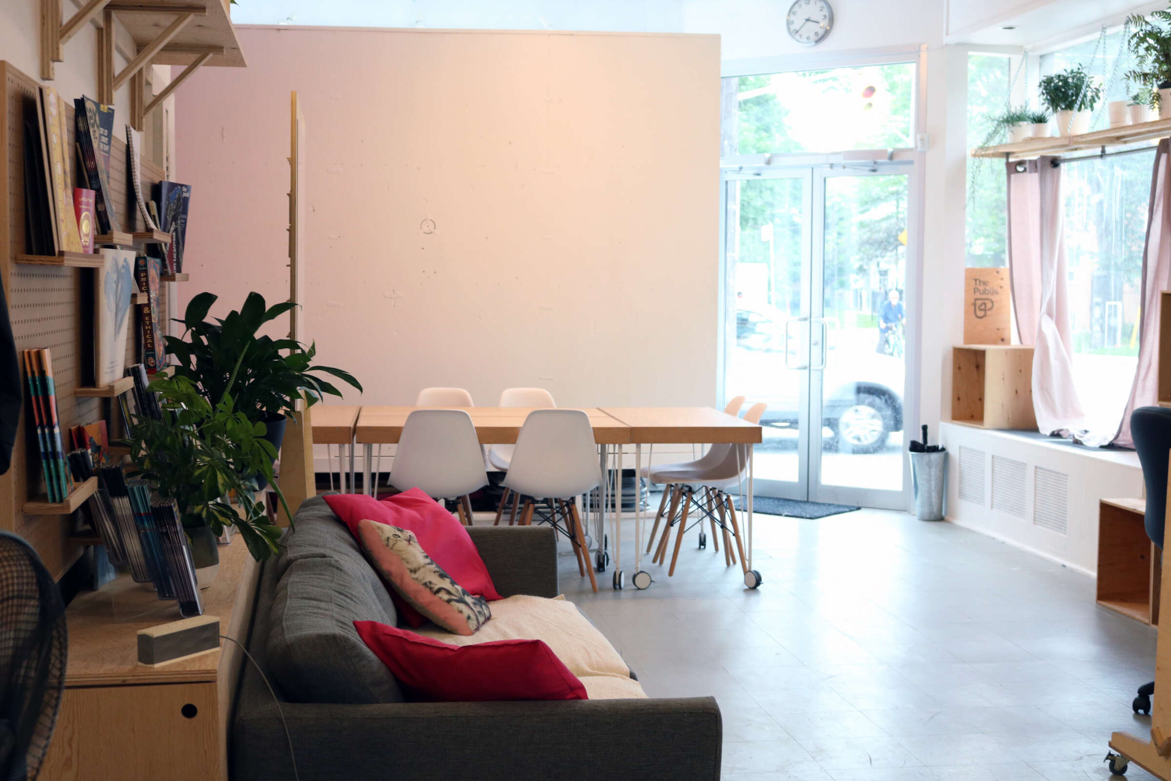 A cozy, well-lit room with a grey couch adorned with red and beige pillows, a wooden table surrounded by white chairs, and shelves filled with books and plants. Large windows let in natural light, and a door leads outside.