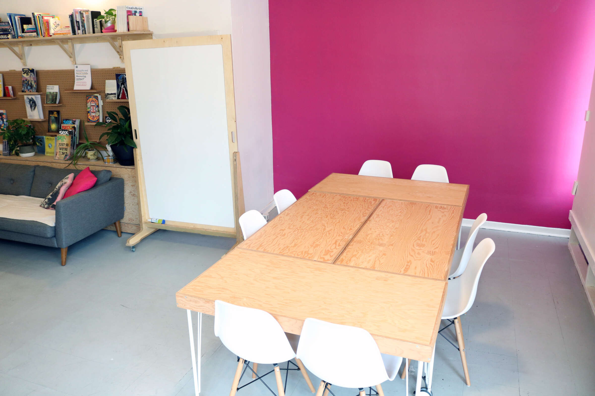 A meeting room with a wooden table surrounded by six white chairs. In the background, there's a couch with a pink and a turquoise pillow. A bookshelf full of books and plants lines the wall. One wall is bright pink, adding a vibrant touch to the space.