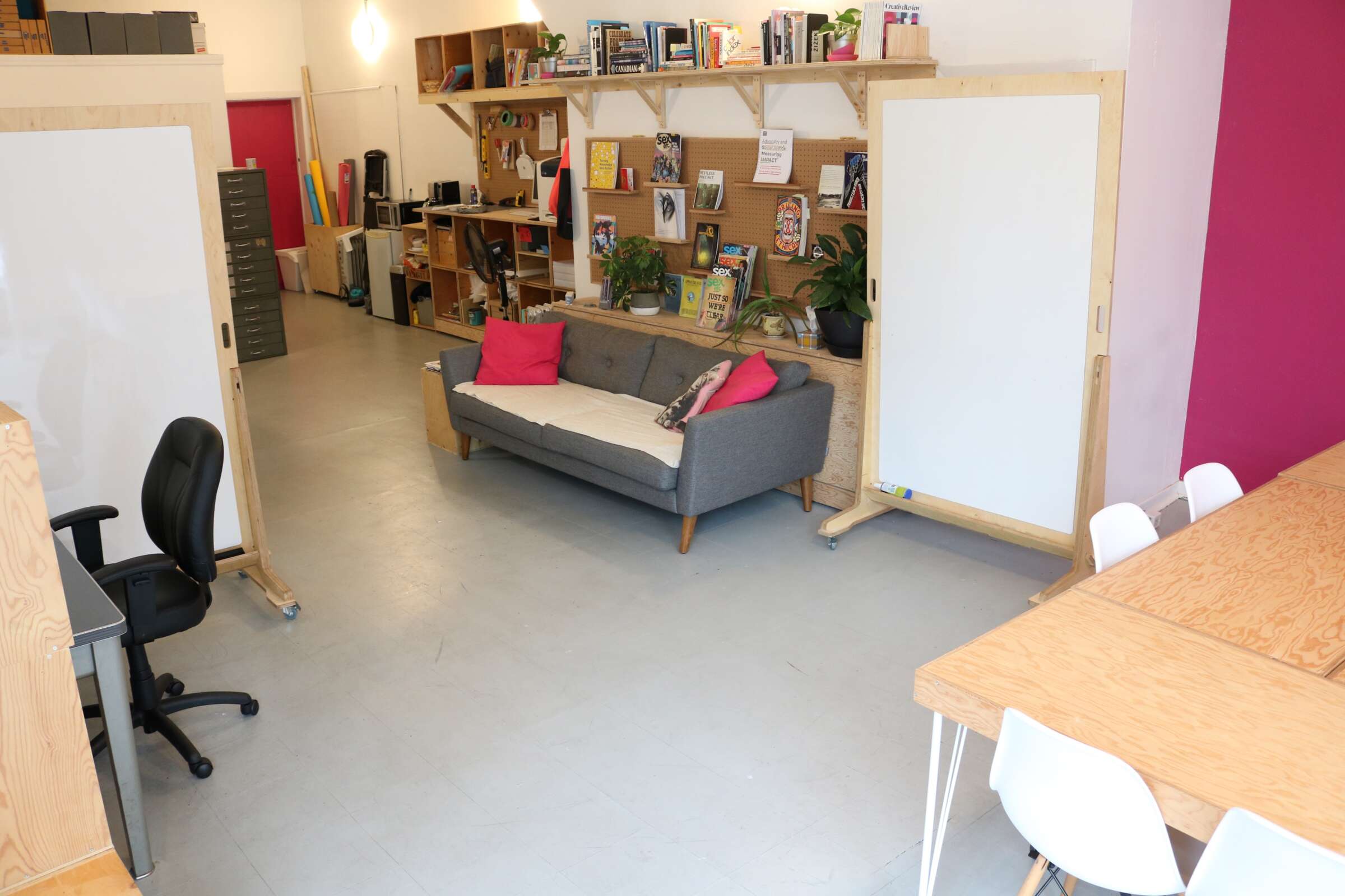 A modern office space with a gray couch adorned with pink and white cushions. A bookshelf filled with books and decorations is behind the couch. There are whiteboards, a desk with chairs to the right, and an office chair in the foreground. The floor is light gray.