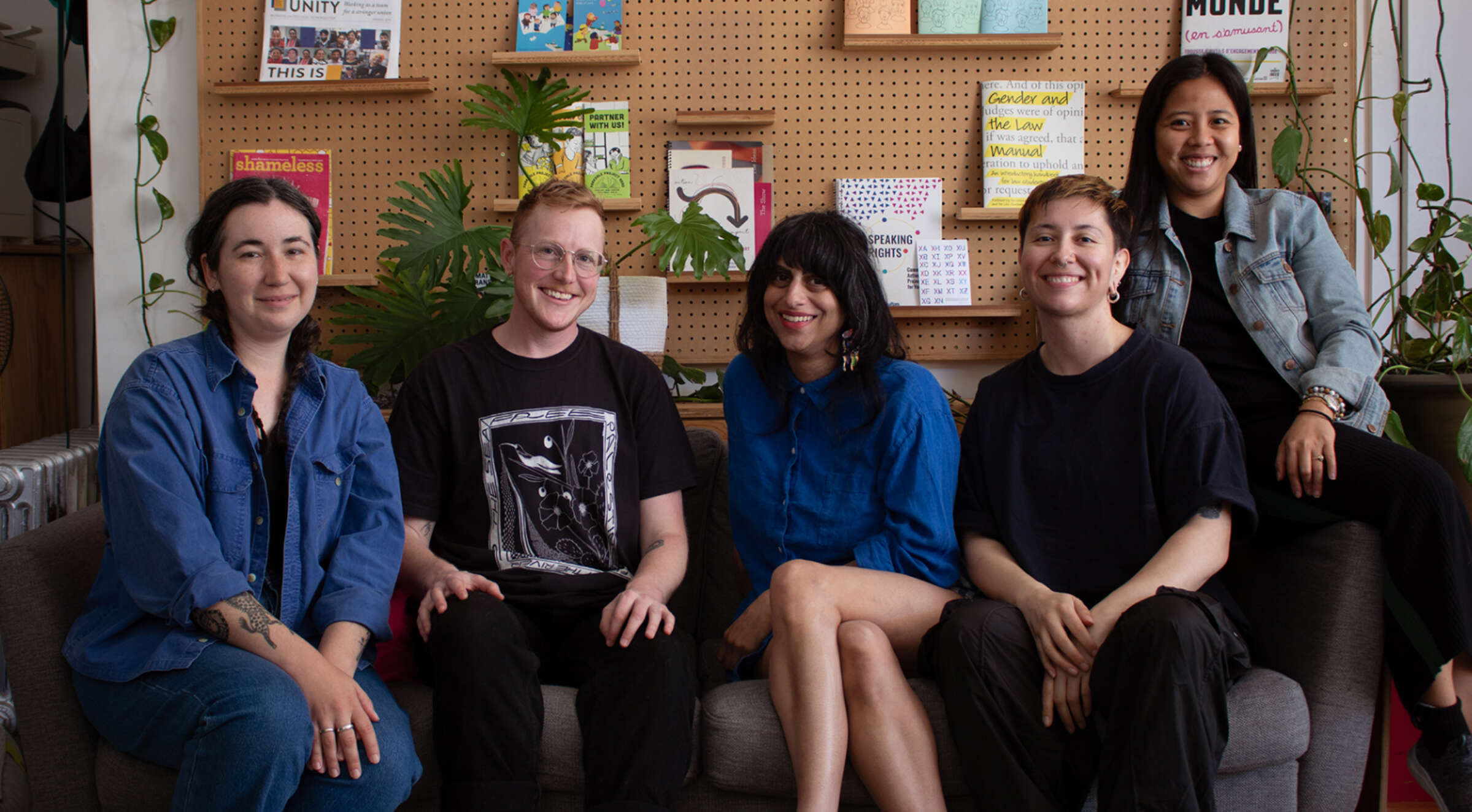 A group of five people sits on a sofa, smiling for the camera. Behind them is a pegboard wall with books, plants, and various items on display. The setting appears cozy and creative, with greenery and warm lighting contributing to a welcoming atmosphere.
