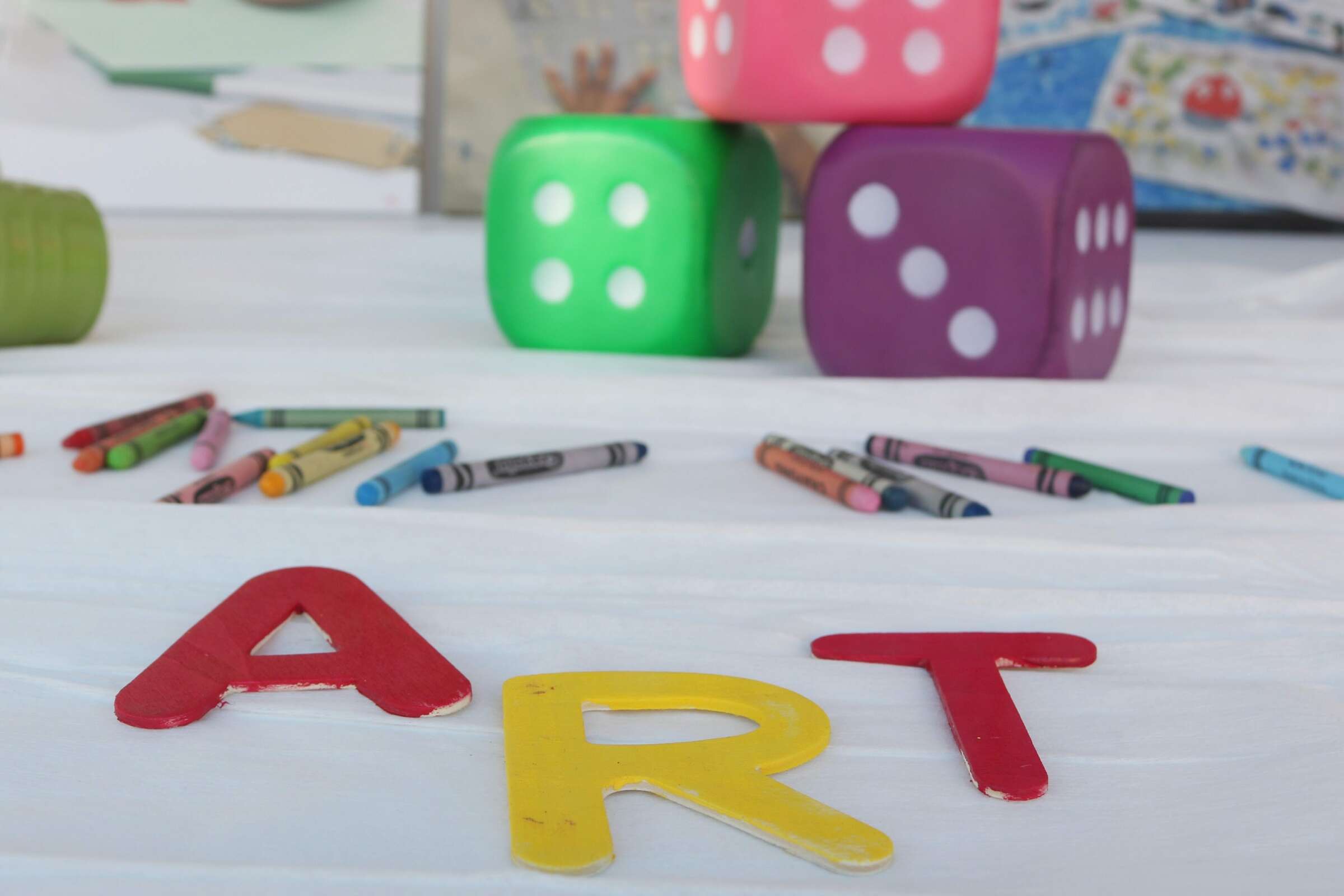 Several crayons lie scattered on a white surface, with three large dice and colorful letter cutouts spelling "ART" in the foreground. An assortment of creative supplies and artwork is visible in the background.