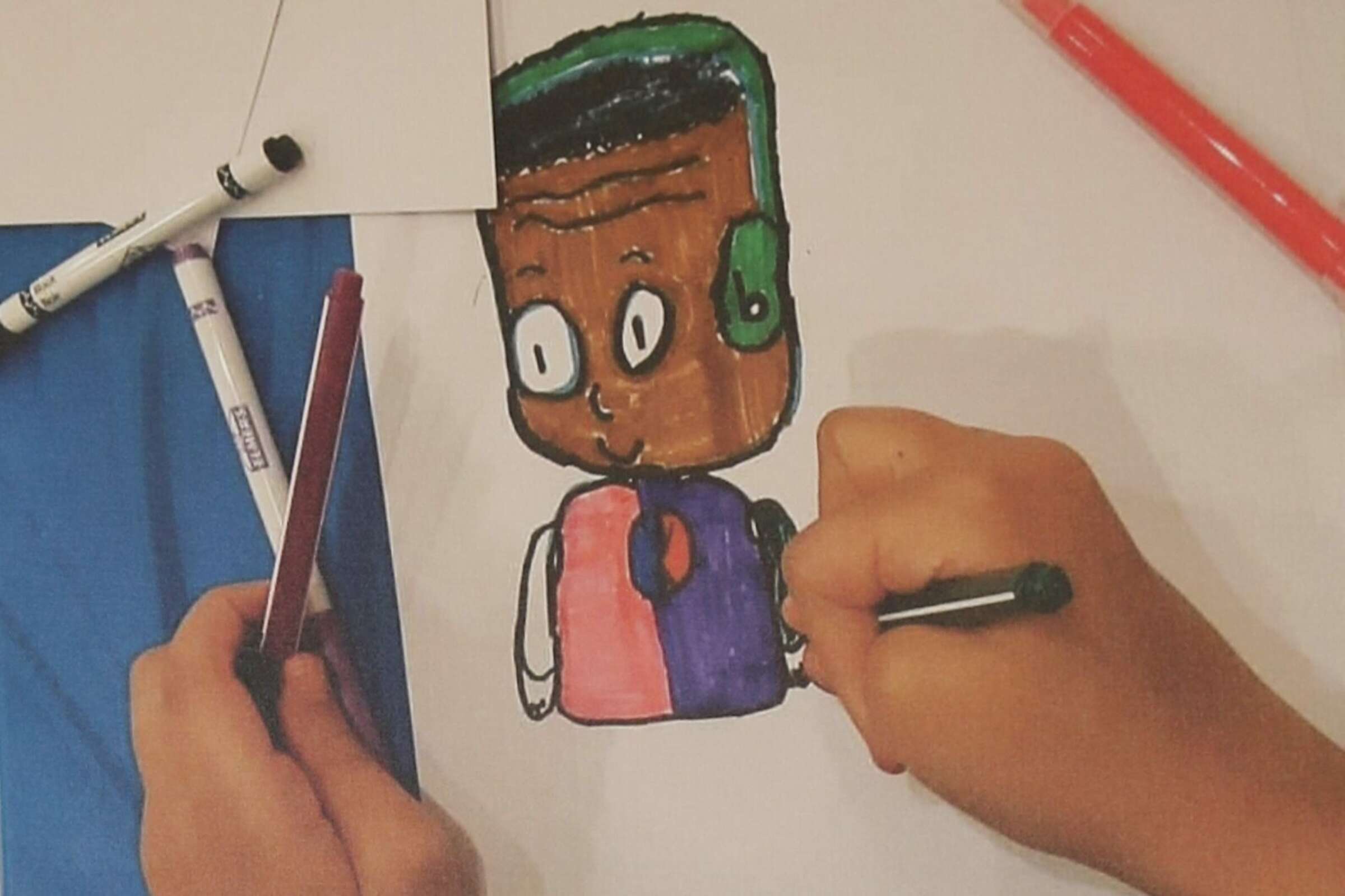 A child's hands are seen coloring in a drawing of a cartoon character on white paper. The character has green hair and a brown face, with a pink and blue outfit. Various colored markers, including red and black, are scattered around the drawing area.