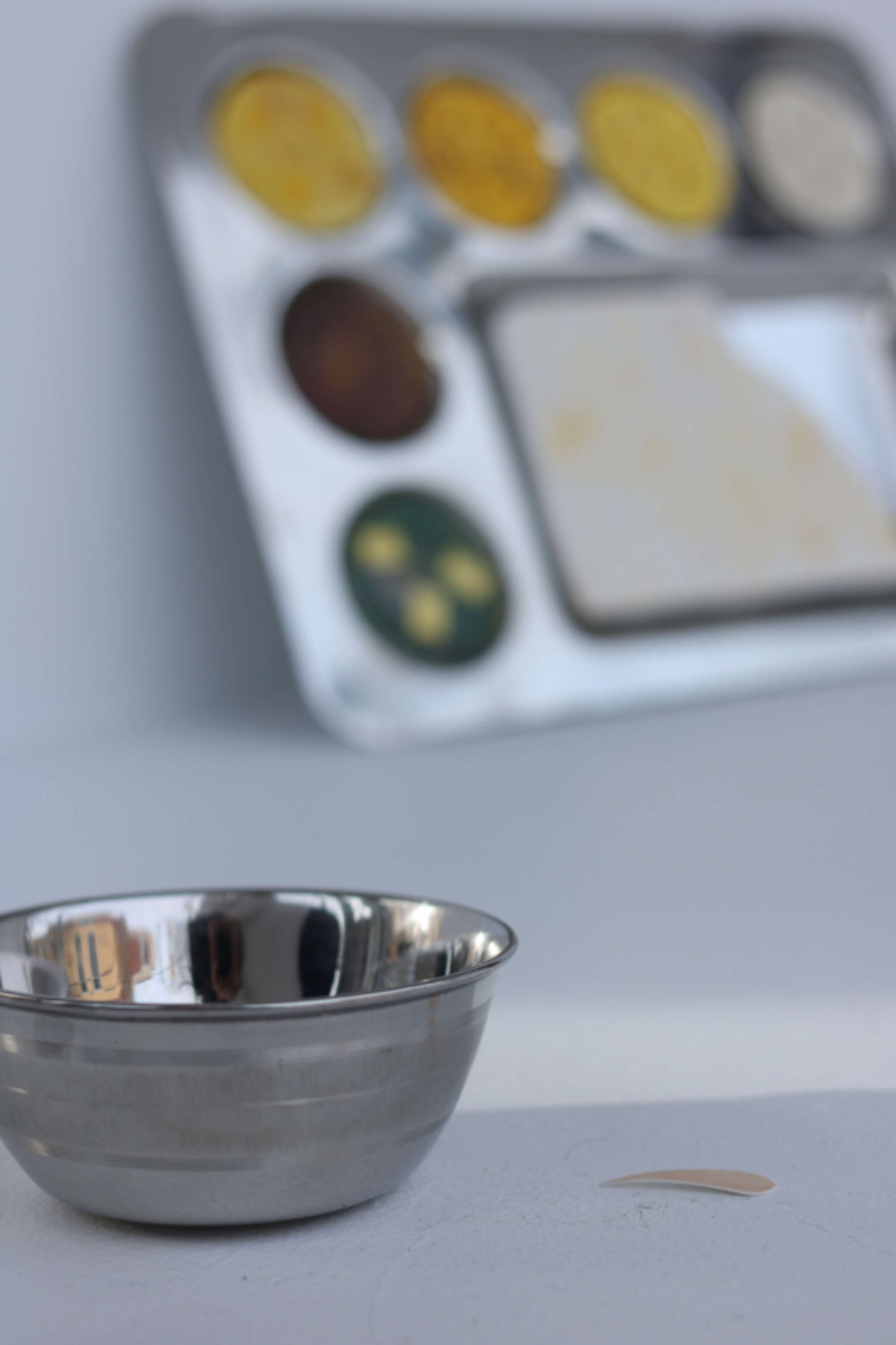 A small stainless steel bowl sits on a light-colored surface with a tiny adhesive bandage beside it. In the blurred background, there is a metal tray holding several paint wells with yellow, brown, and green paint.