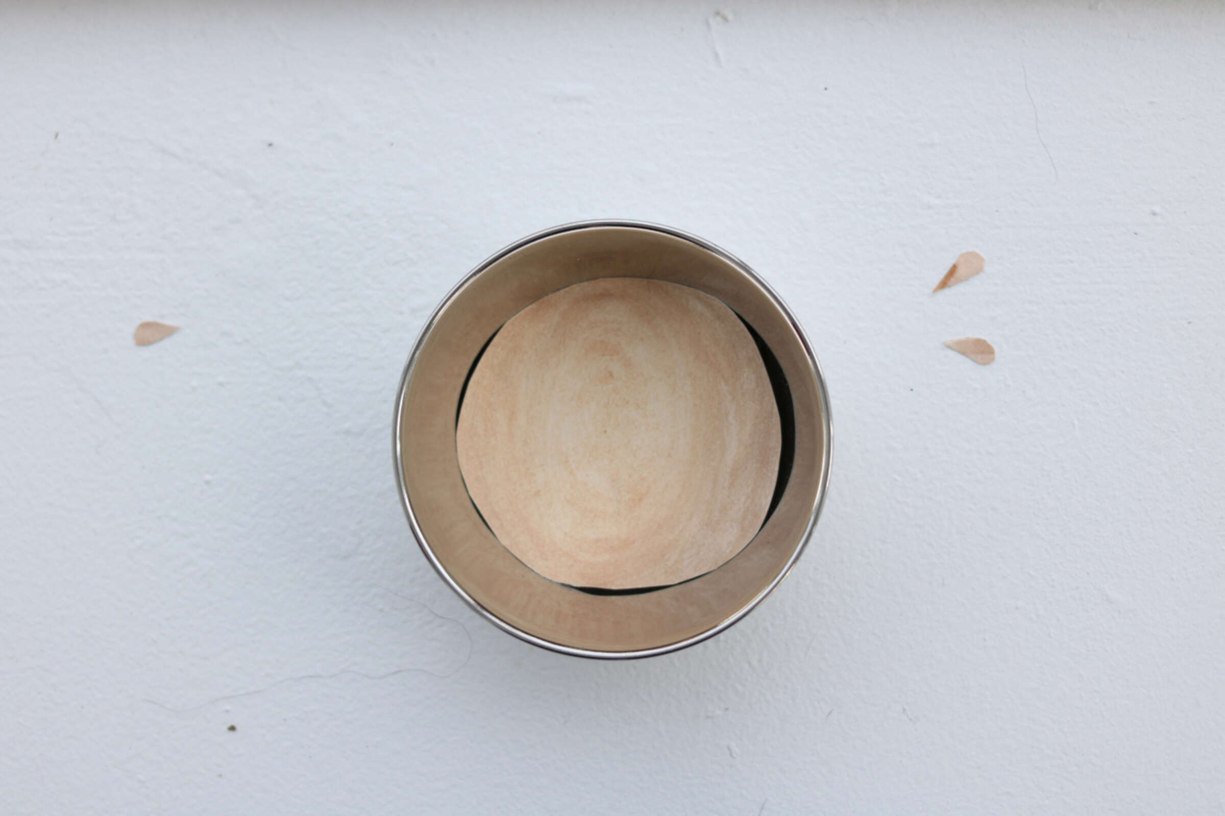 A round, empty metal container with a wooden base, viewed from above, is placed on a white surface. Two small teardrop-shaped marks are visible on the surface near the container, suggesting a minimalist or abstract design.