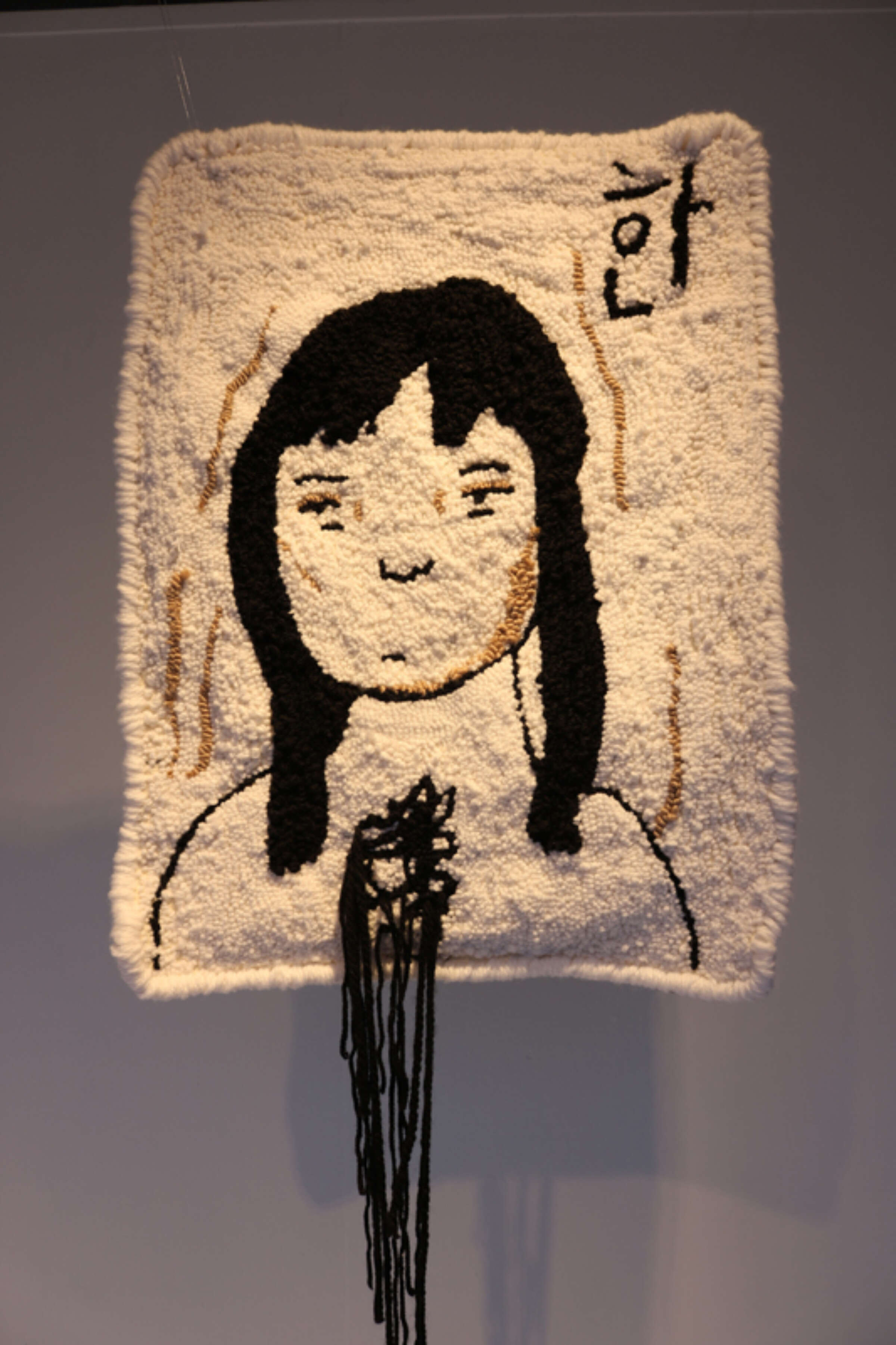 A woven tapestry featuring a simple illustration of a person with long black hair and closed eyes. The person's hands are clasped together in the center. The tapestry has an off-white background with minimalist lines and black strings hanging from the bottom.