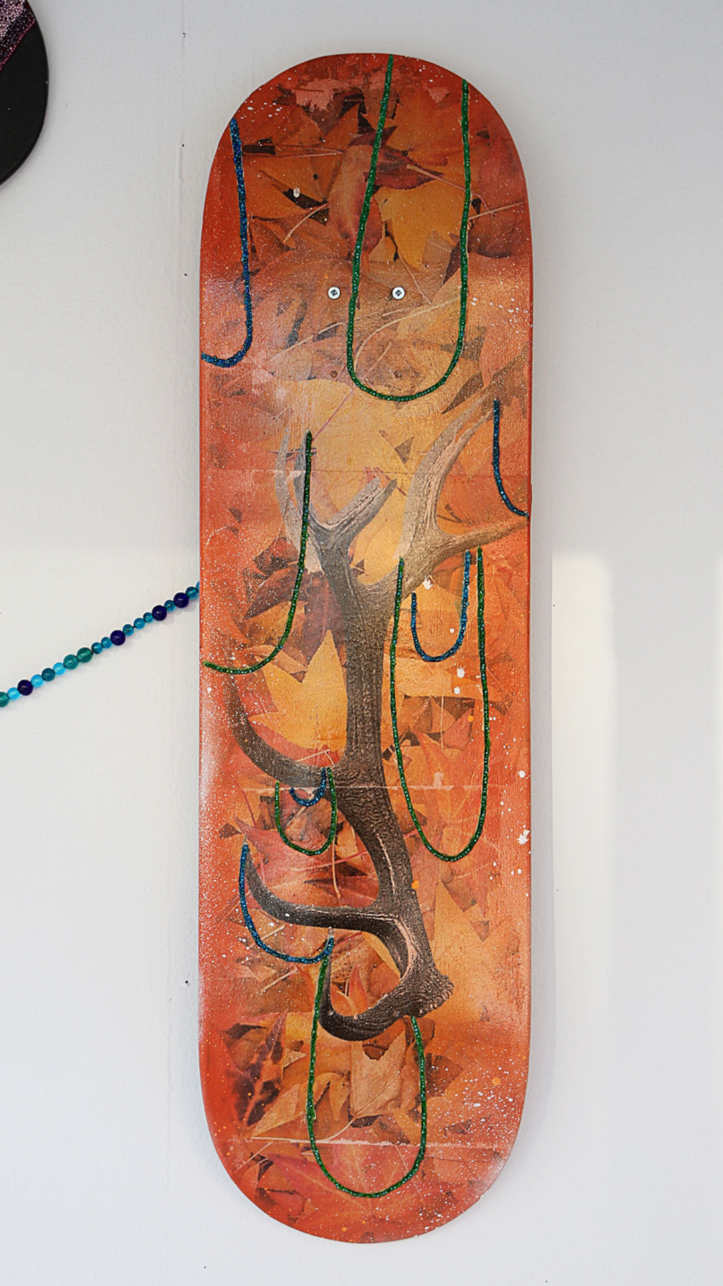 A skateboard deck decorated with autumn leaves and an antler design, painted in vibrant orange, brown, and green. The antlers are prominently featured, with green outlines around them. The deck is displayed vertically on a white wall.