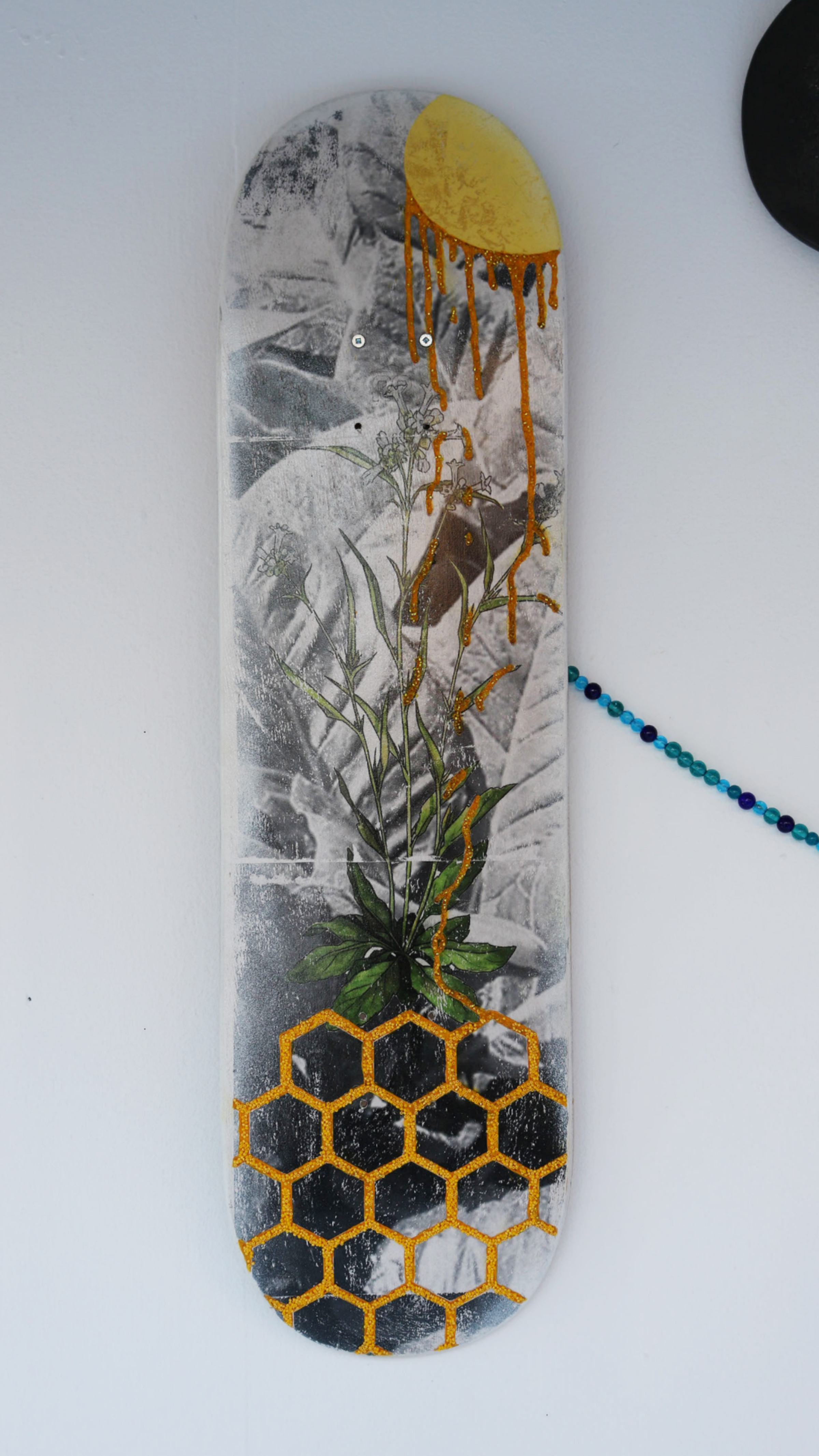 A skateboard deck with a mixed media design featuring golden honeycomb patterns at the bottom, a green plant in the middle, and a partial yellow sun dripping paint at the top against a monochrome background. A string of blue and purple beads hangs on the side.