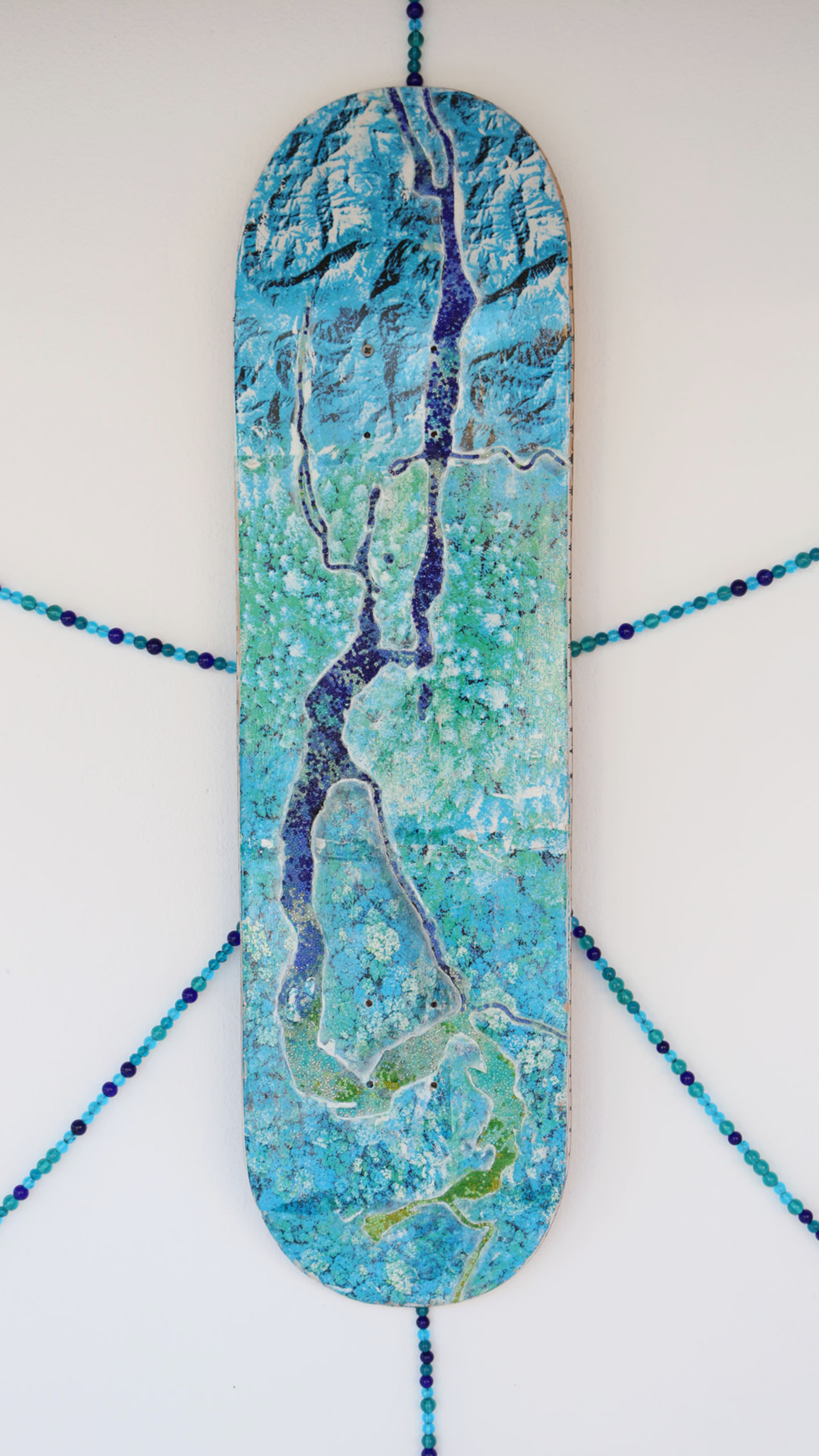 A vertical, painted skateboard deck mounted on a white wall, featuring a blue and green abstract design mimicking a topographic map. Surrounding the skateboard is a symmetrical arrangement of blue and purple beads extending outward like rays.