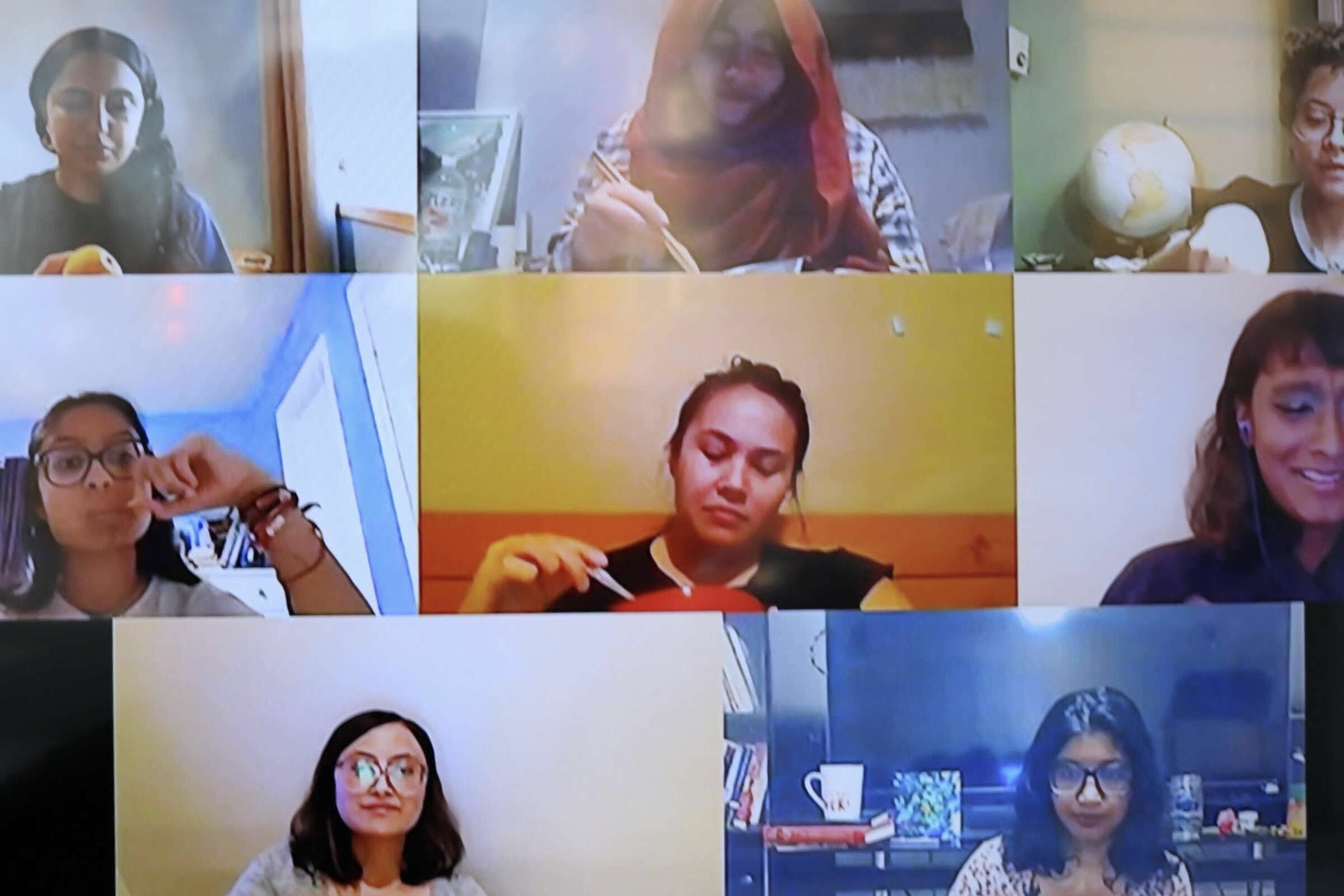 A screenshot of a video call shows nine women in separate boxes, each engaged in different activities. Some are eating, one is holding a globe, and another is writing. They appear to be in a meeting or casual group conversation.