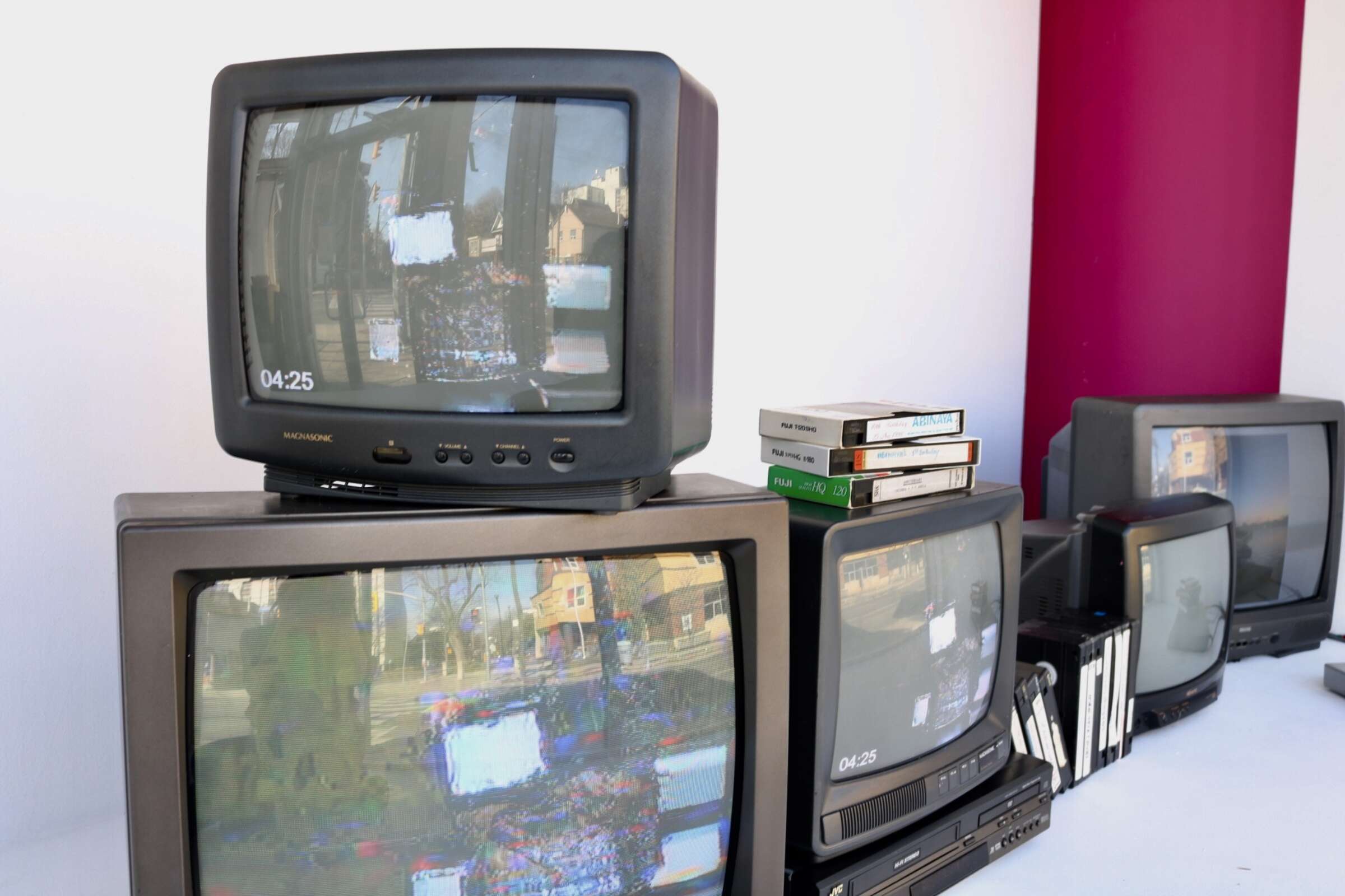 Several vintage CRT televisions are arranged together, most displaying distorted images with static. Three VHS tapes and a few books are stacked on top of one of the TVs. A magenta and white wall provides a colorful backdrop. The time "04:25" is visible on two screens.