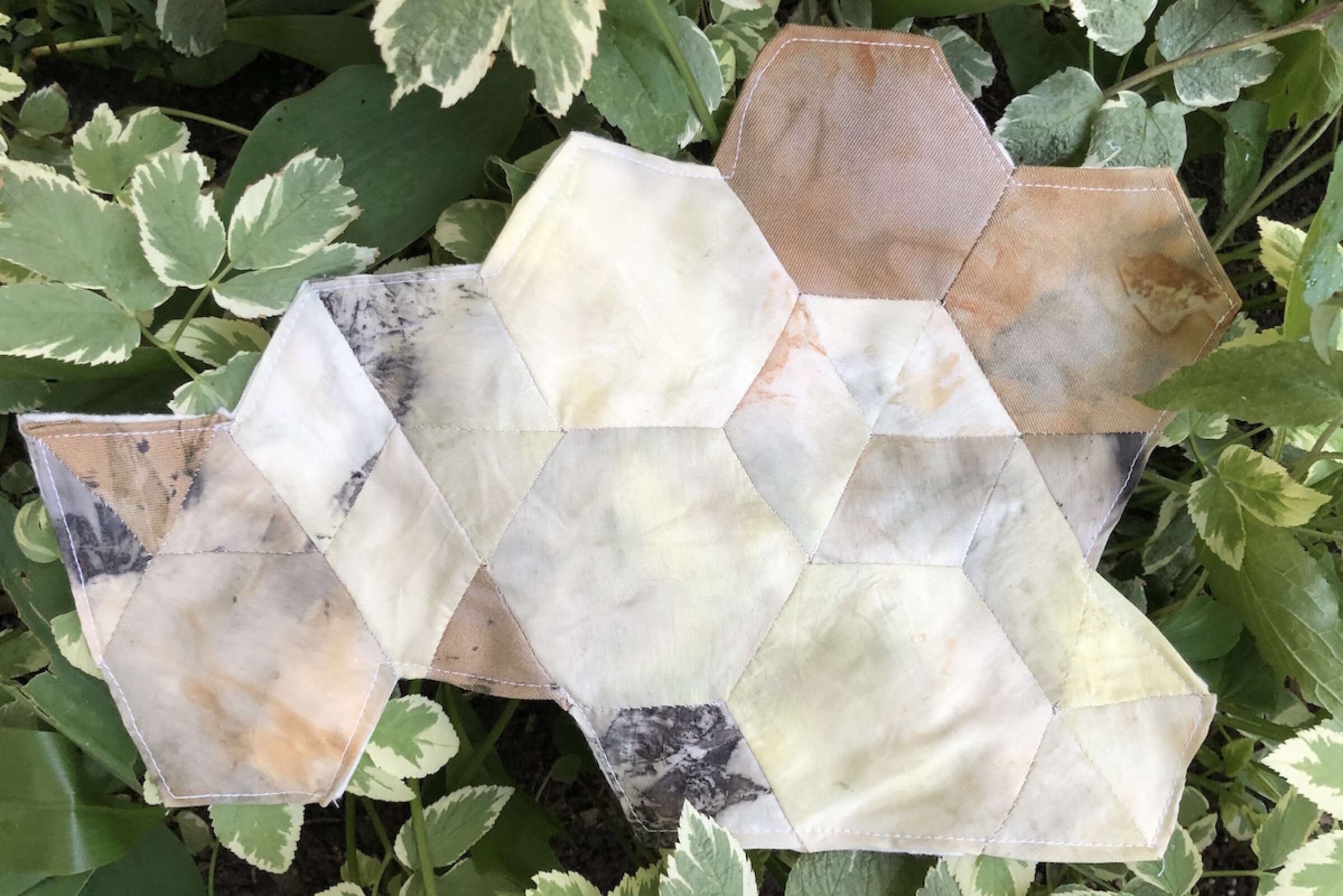 A handmade geometric patchwork, consisting of hexagon and pentagon shapes in various shades of beige, brown, and cream, is displayed against a backdrop of green variegated plants. The patchwork appears to be made from fabric pieces stitched together.