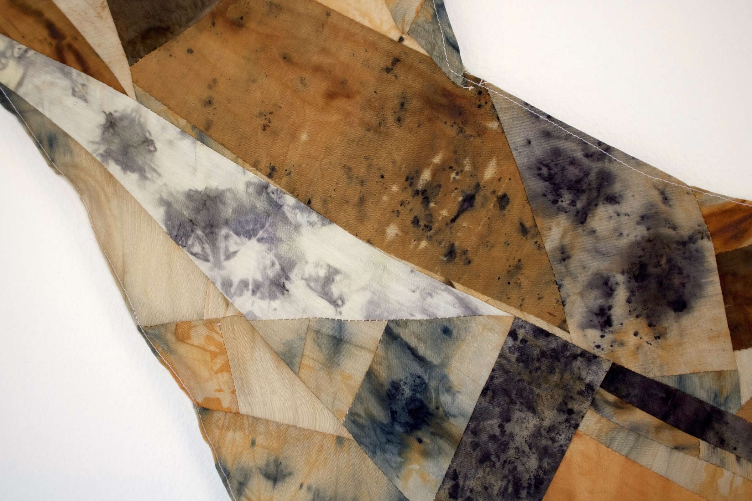 An abstract textile artwork featuring irregular geometric shapes in earthy tones like tan, beige, and brown with areas of dark blue and gray. The fabric appears to be hand-dyed, showing intricate, organic patterns and textures.