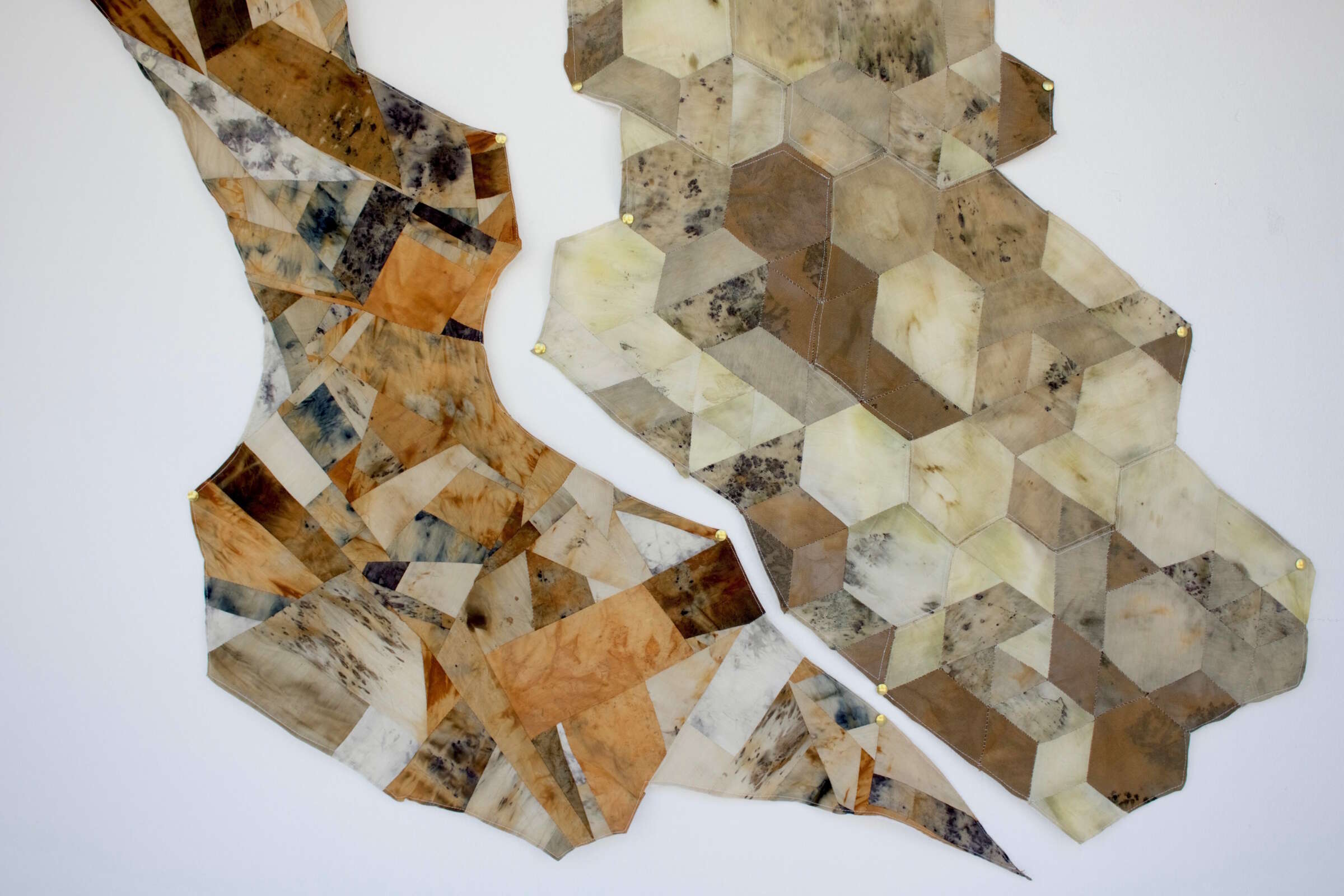 A wall-mounted fabric art piece composed of various geometric shapes and earth-toned colors, including shades of brown, beige, and gray. The artwork is divided into two main sections, each featuring a mix of irregular and hexagonal patterns.