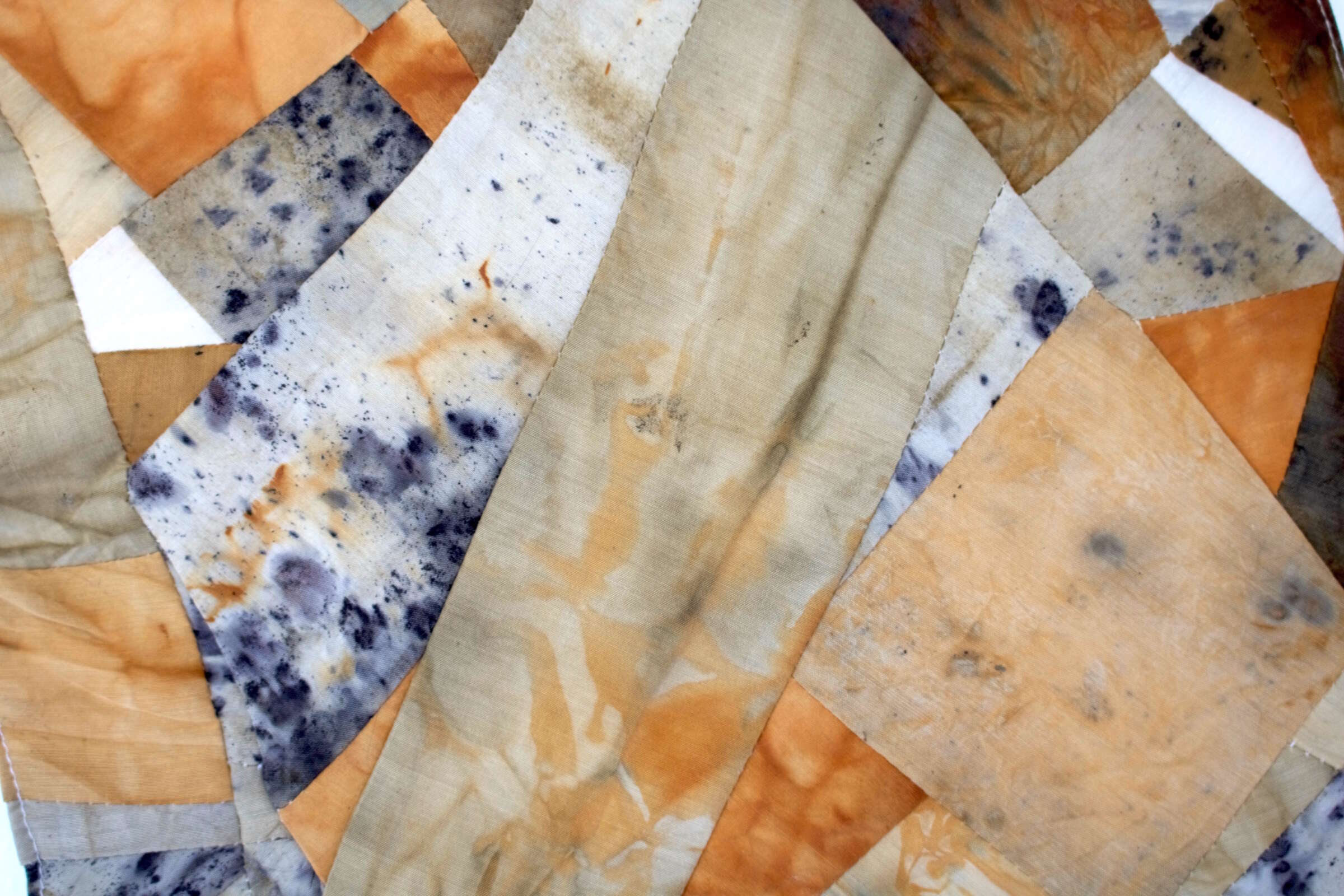 A close-up of a quilt made from fabric patches. The patches vary in colors, including beige, orange, and gray with hints of black spots. The overlapping pieces create a textured, abstract pattern.