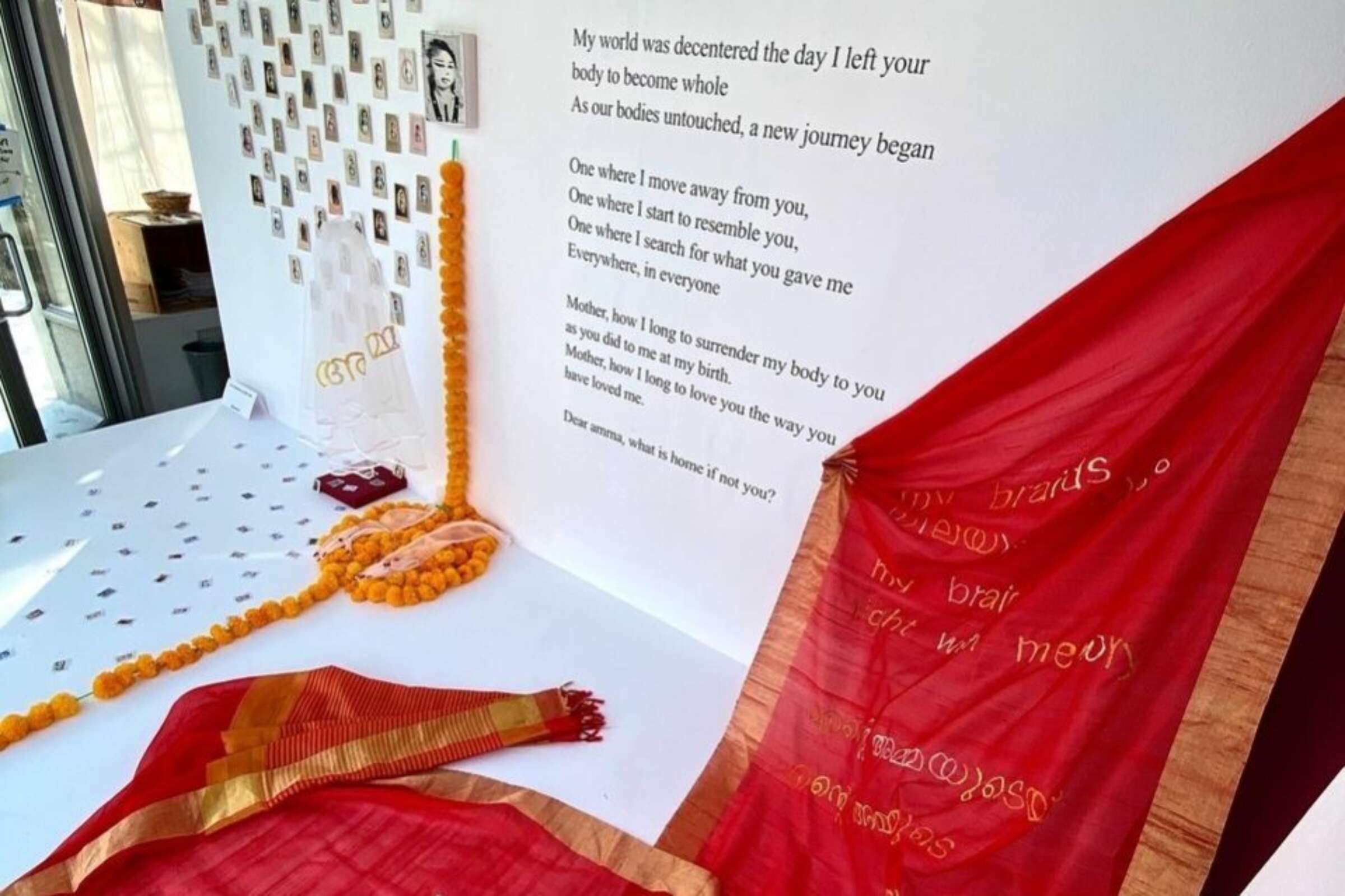 A red sari with gold details is displayed on a table near a white wall adorned with polaroids, yellow flowers, and various items. Text on the wall reads, "My world was decorated the day I left your body to become whole. Our bodies untouched, a new journey began.