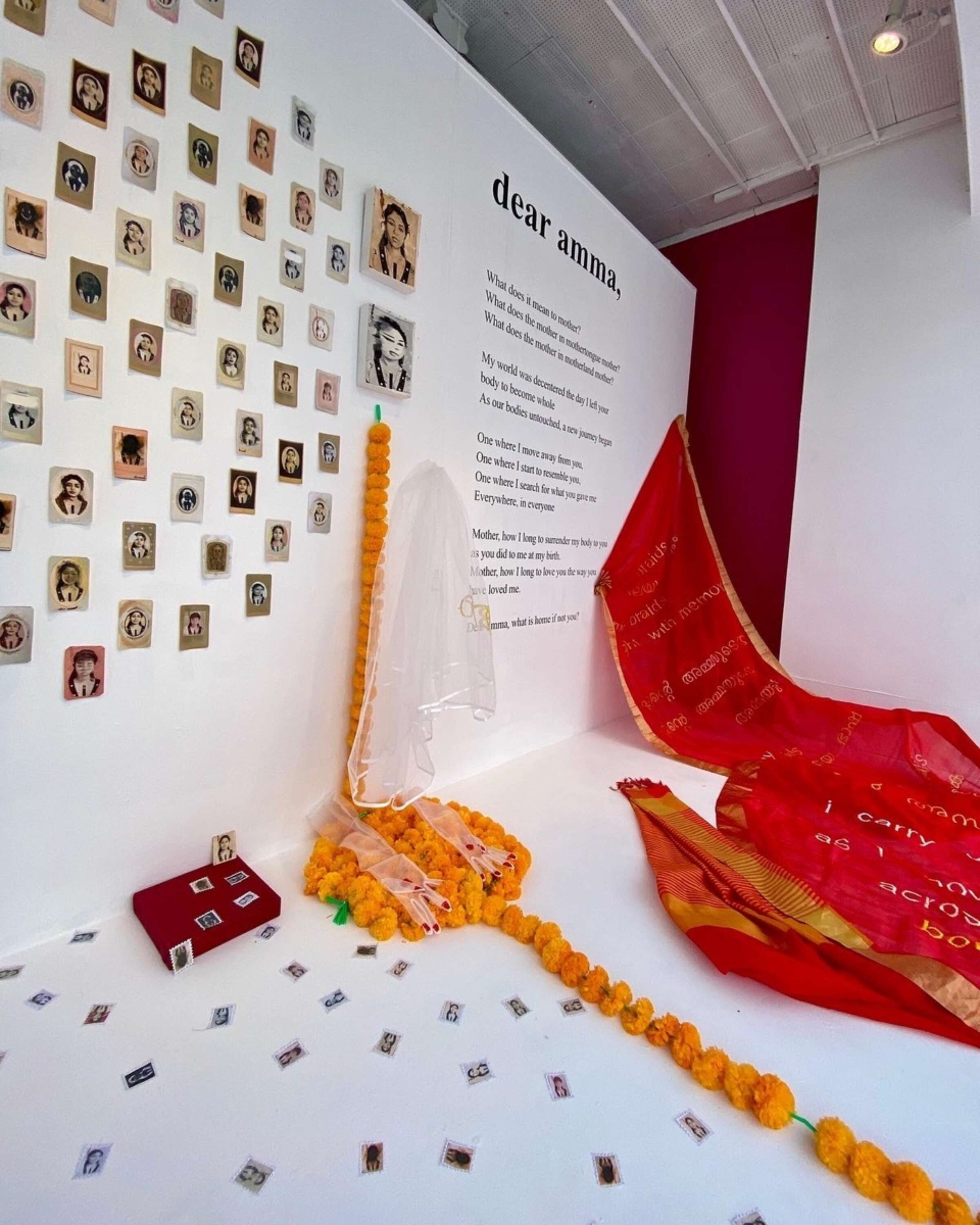 An art installation featuring portraits of women arranged on a white wall, a red fabric draped on the floor, and orange marigold garlands. A large text titled "dear amma," is displayed on the wall. Small photos and objects are scattered on the ground.