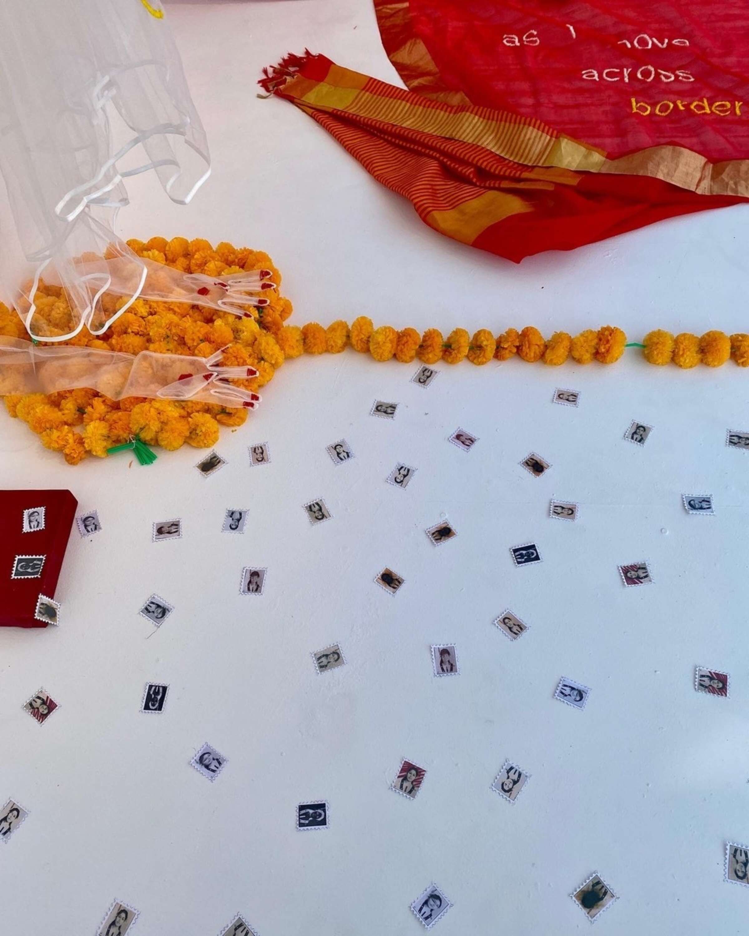 A person's feet are adorned with orange marigold flower garlands on a white floor. Scattered around are small paper squares with black-and-white images. A red sari with gold trim and a visible phrase, "as I love across borde," is partially seen in the background.