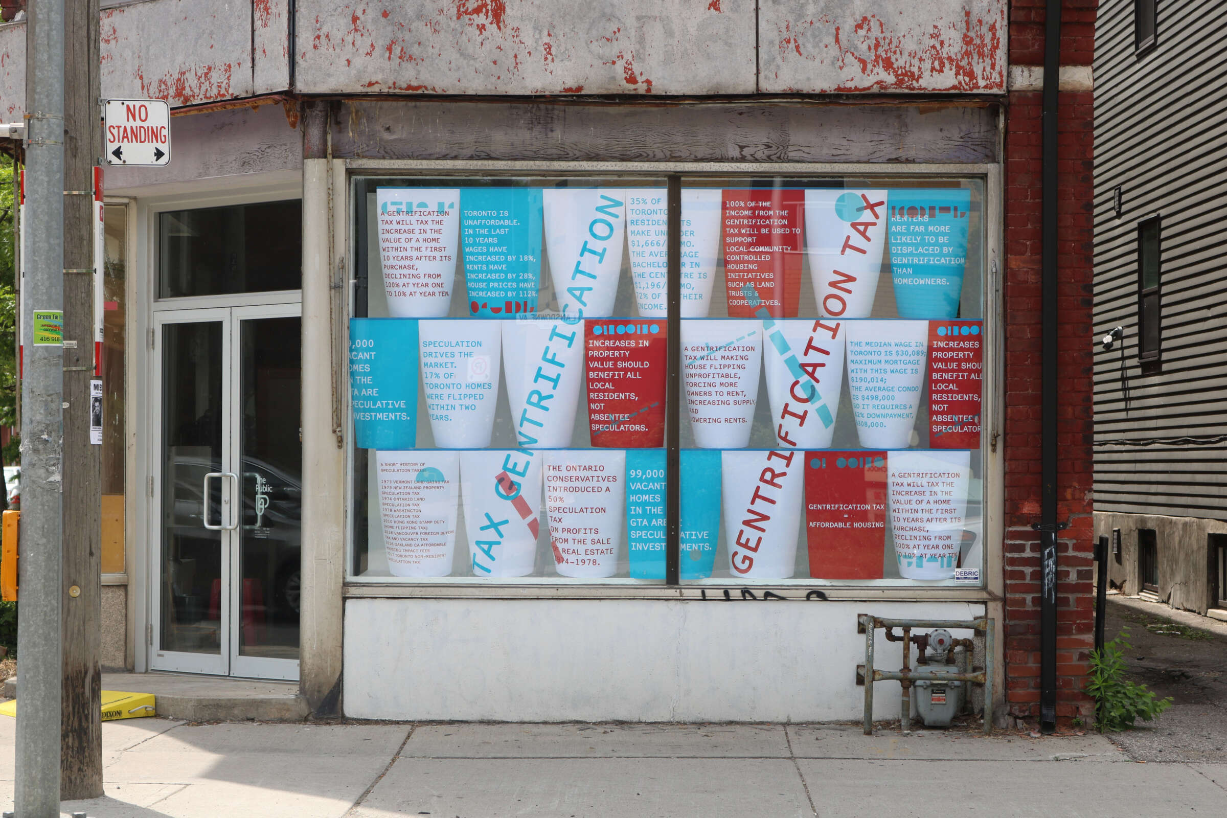 A storefront with a large window covered in colorful posters. The posters have text discussing issues like "gentrification" and "tax incentives." The exterior building is weathered with peeling paint. A "No Standing" sign is mounted on a nearby pole.