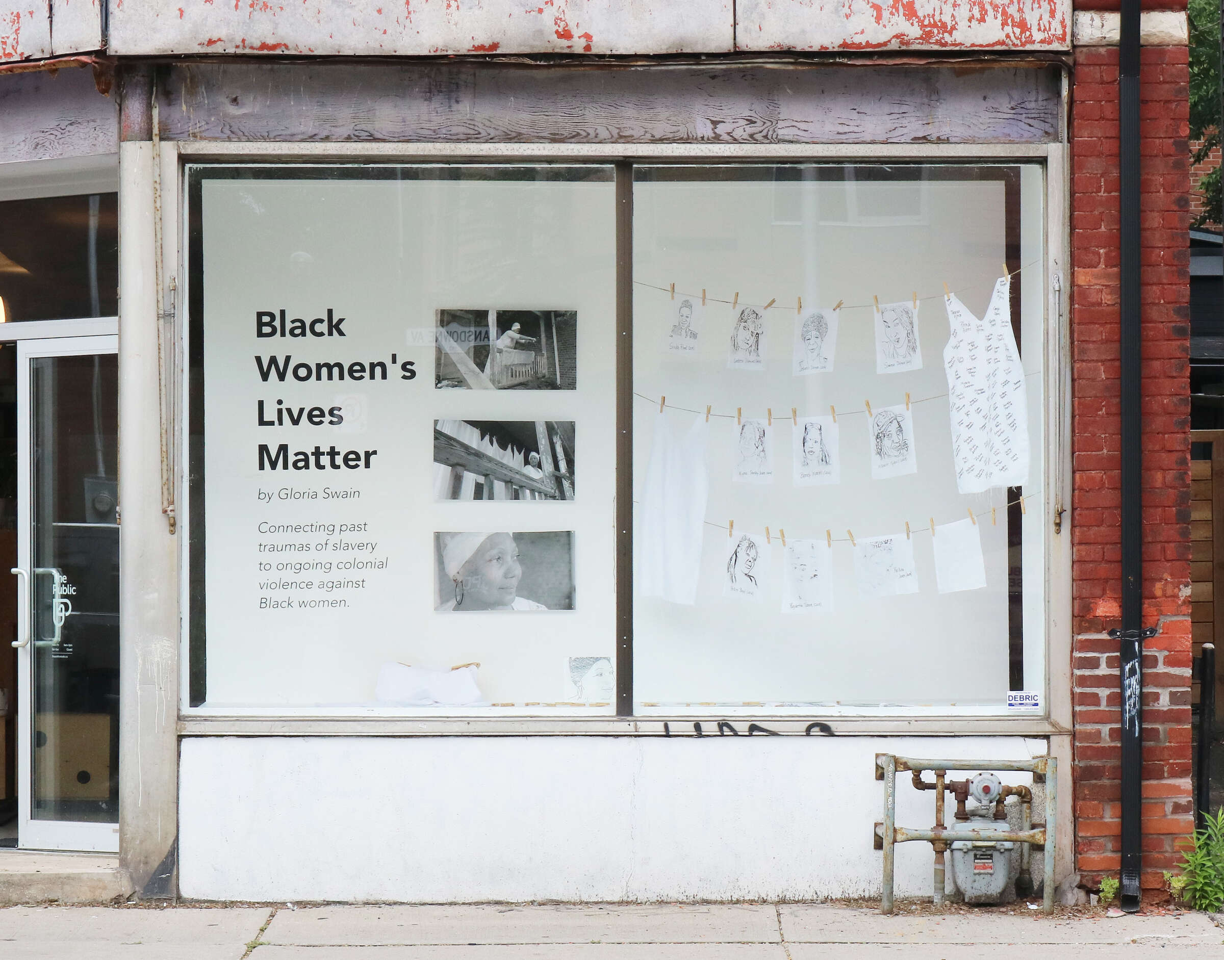 A storefront display features a white wall with the text "Black Women's Lives Matter" by Gloria Swain. Photos and artwork are pinned to strings across the window. The exterior shows an aged, red brick building and part of a sidewalk.