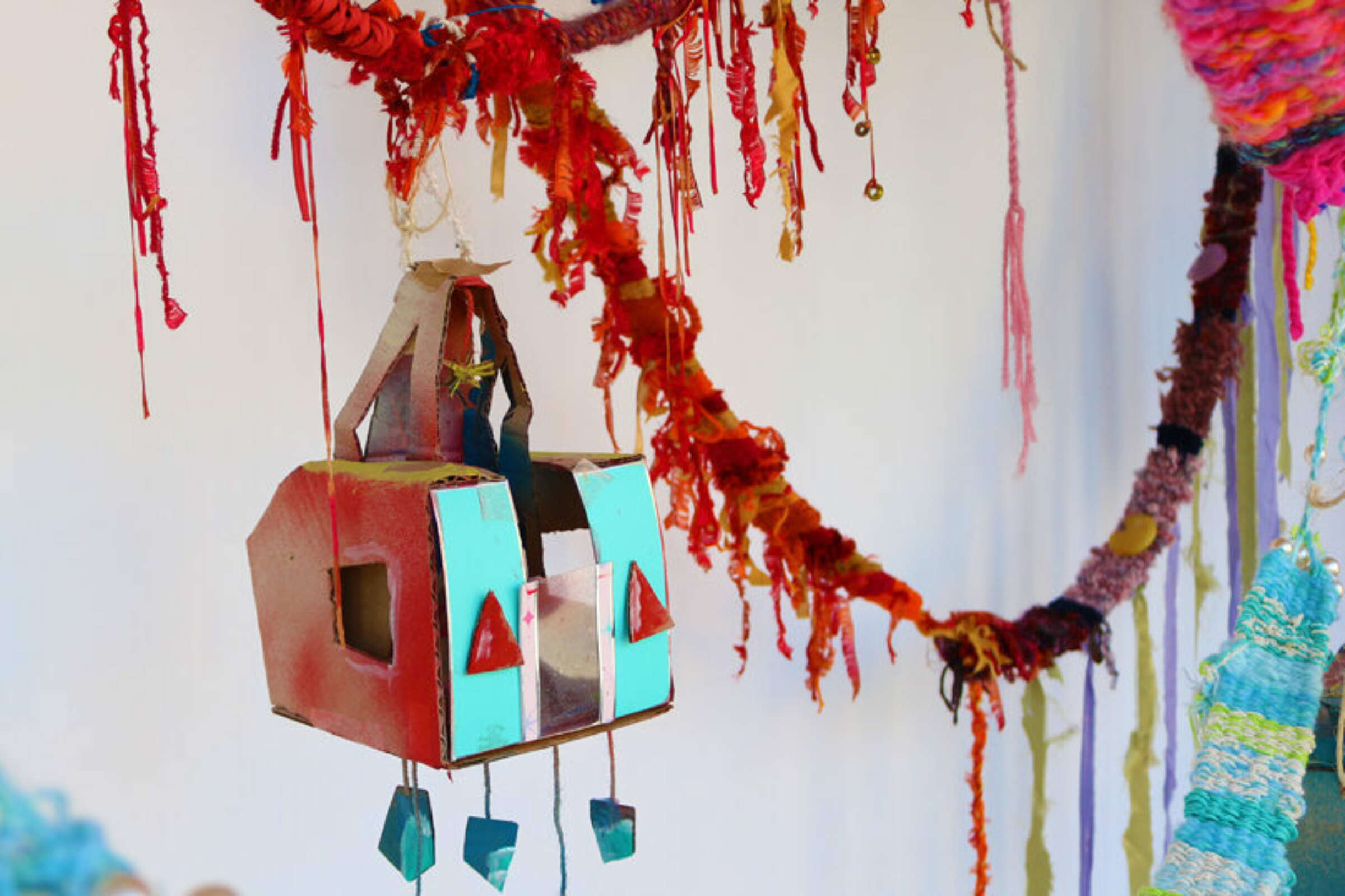 A whimsical homemade birdhouse crafted from cardboard, painted in red and turquoise, is decorated with triangle shapes. It hangs among a variety of colorful yarn and string decorations, creating a vibrant and eclectic backdrop.