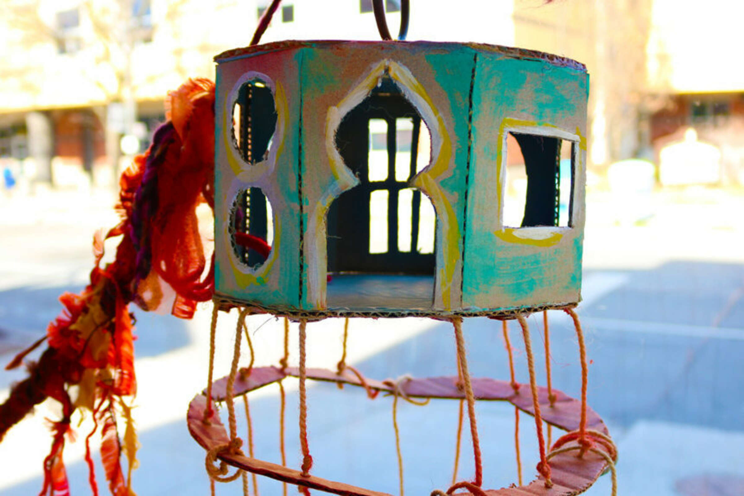 A close-up of a handcrafted, colorful birdhouse with intricate cutout designs, adorned with red and orange fabric strips and suspended from twine, hangs in front of a blurred urban background. Sunlight illuminates the detailed artwork, showcasing its vibrant hues.