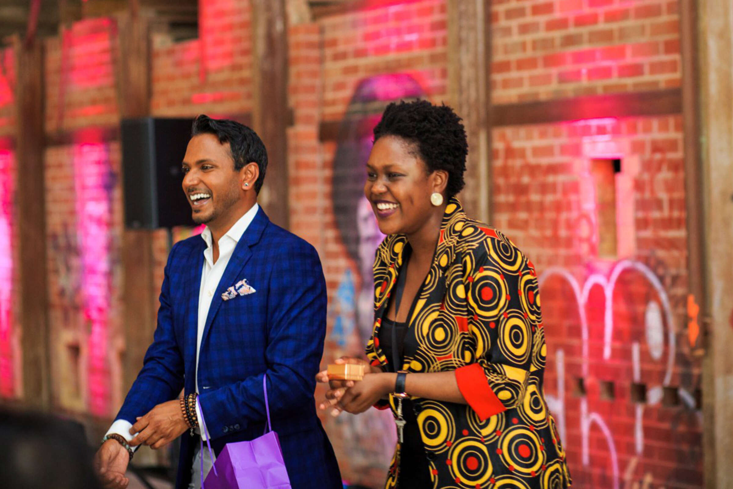 Two people stand in front of a brick wall with colorful lighting, smiling and engaged in conversation. The man on the left wears a blue checkered suit, and the woman on the right wears a vibrant patterned jacket. The atmosphere appears joyful and lively.